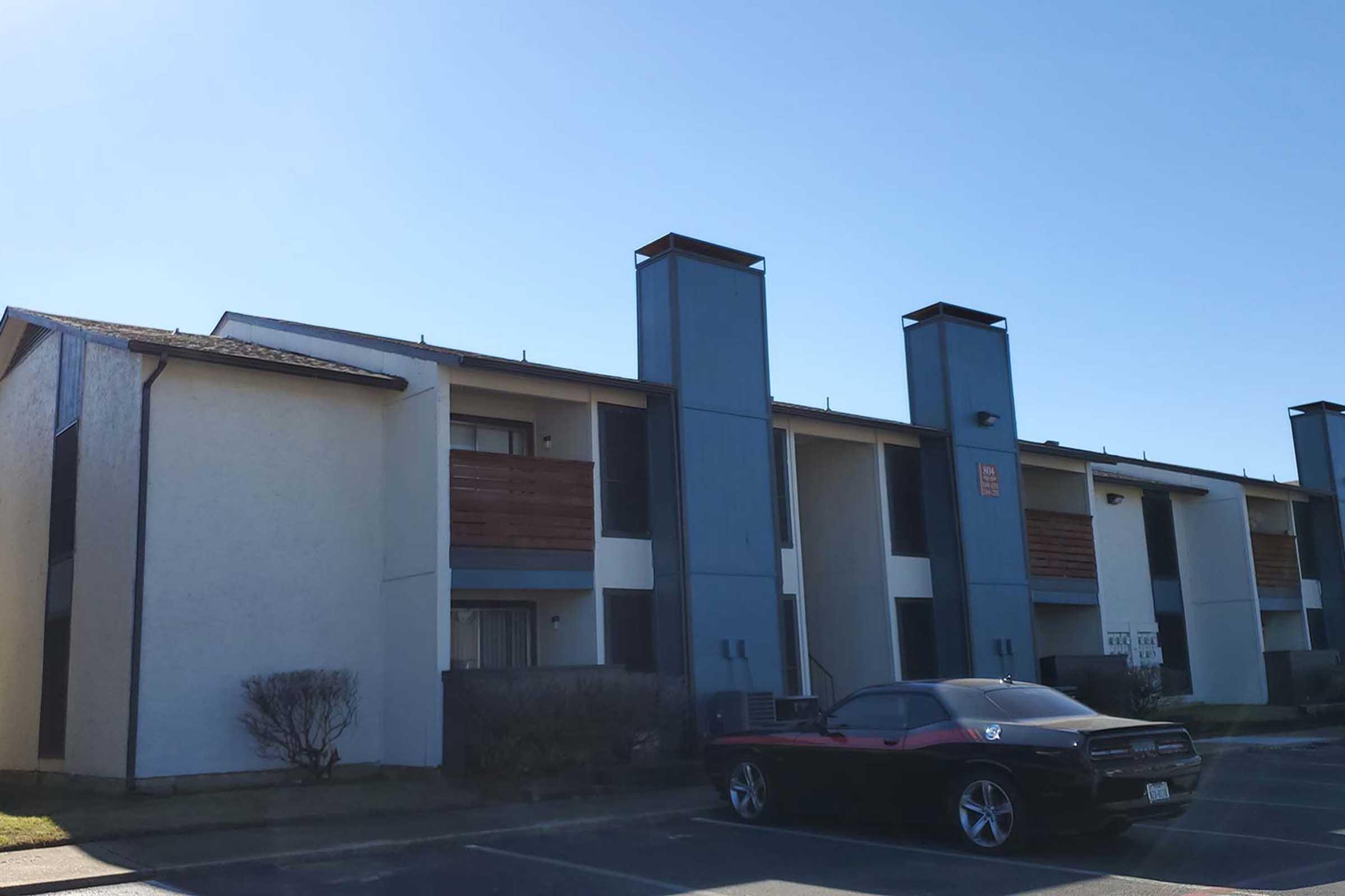 A two-story apartment building with a light-colored exterior and wooden balconies. The building features tall, blue chimneys on the roof. In the foreground, a black car is parked in the lot. The sky is clear and sunny, indicating a bright day.