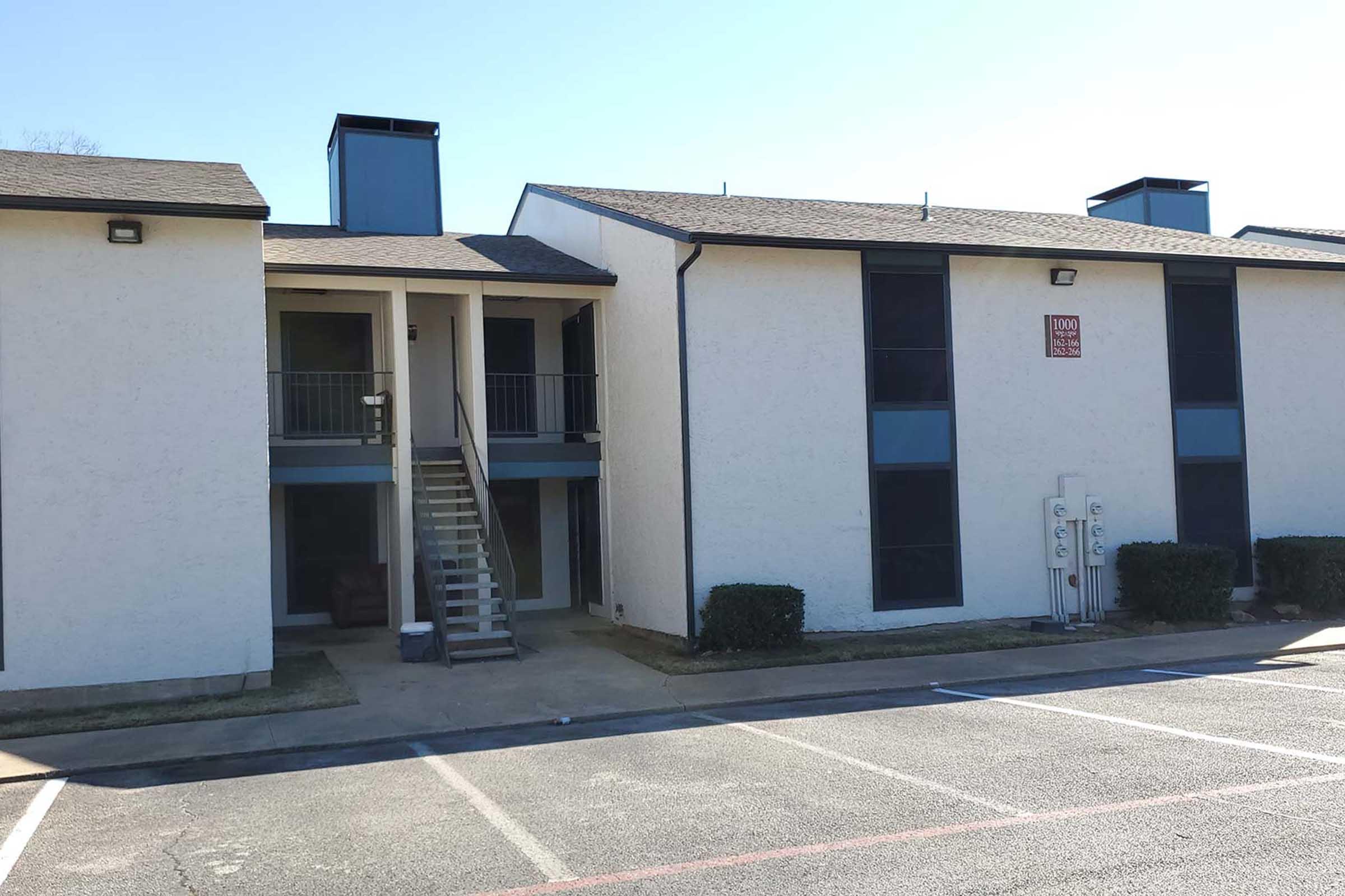 Two-story apartment building with a light-colored exterior, featuring a central staircase leading to upper units. The ground level has utility meters visible, and the area is surrounded by small hedges. The parking lot is empty, and clear blue sky is visible above the building.
