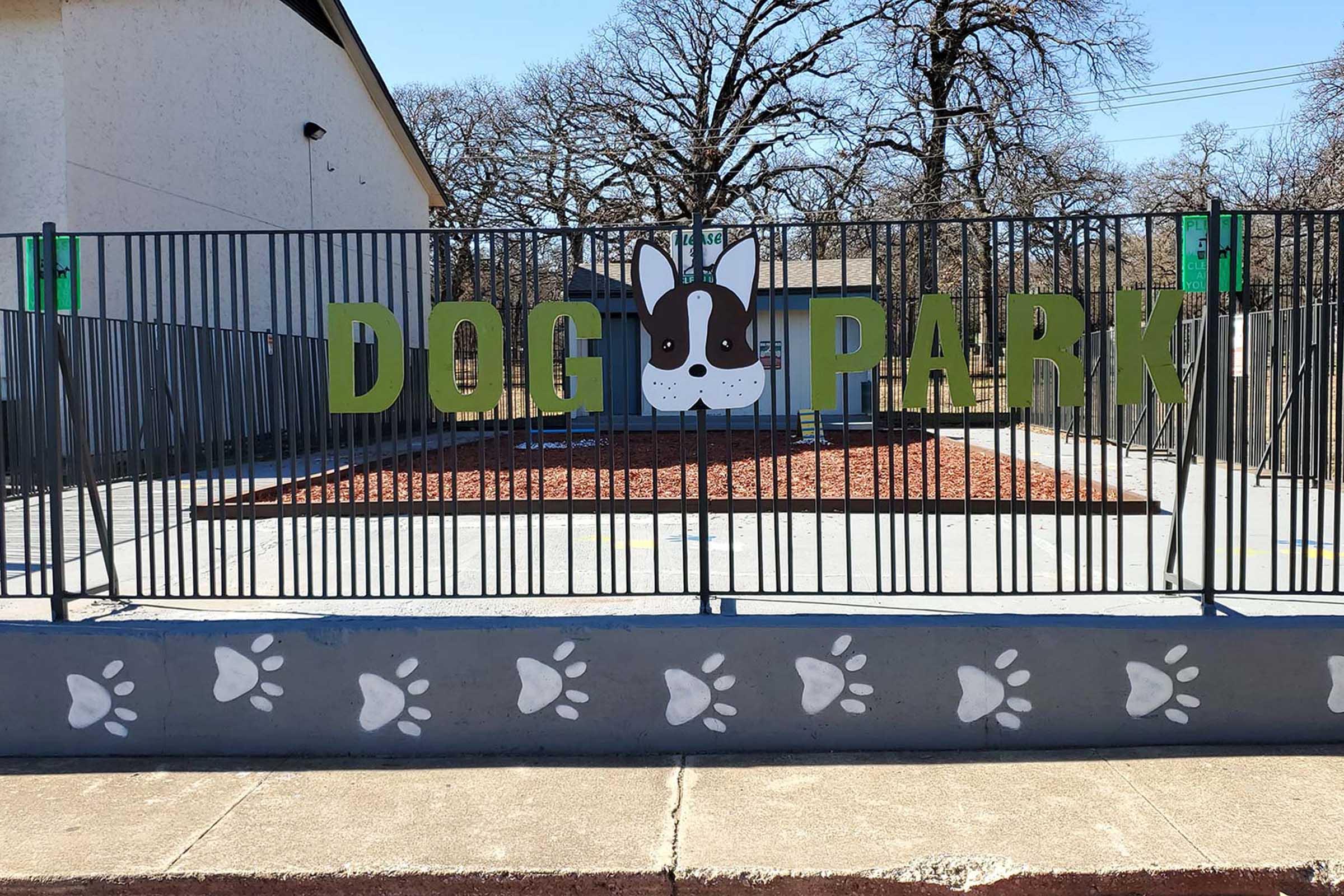 A fenced dog park with a large sign that reads "DOG PARK." The sign features a cartoon dog face in the center. The ground inside the park is covered with mulch, and the fence has decorative paw print designs at the bottom. Trees are visible in the background, indicating an outdoor setting.