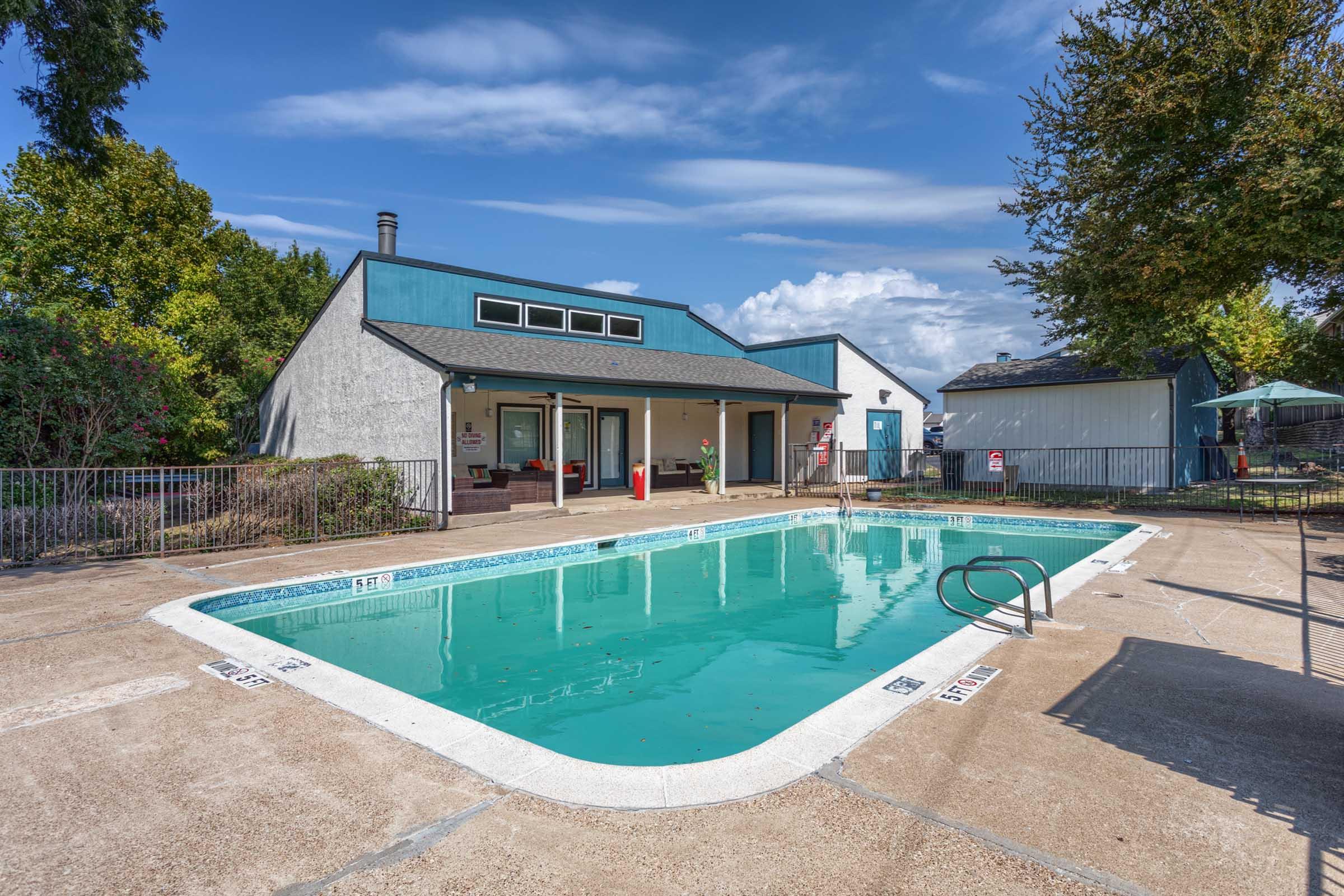 A swimming pool surrounded by a concrete deck, featuring a ladder for entry. In the background, there is a building with a modern design and large windows. Lush greenery and trees are present around the pool area, with a clear blue sky and fluffy clouds overhead.