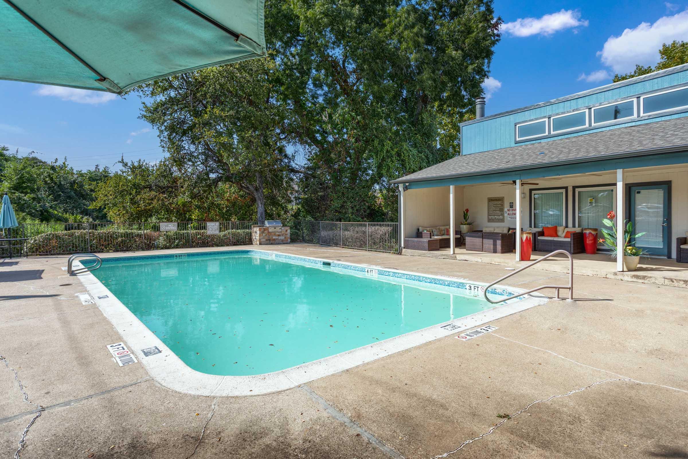 A clear blue swimming pool surrounded by concrete, with a metal railing on one side. There are a few leaves on the water's surface. Nearby, a shaded area with an umbrella and seating is visible, along with a building featuring large windows and an outdoor seating area. The sky is partly cloudy.