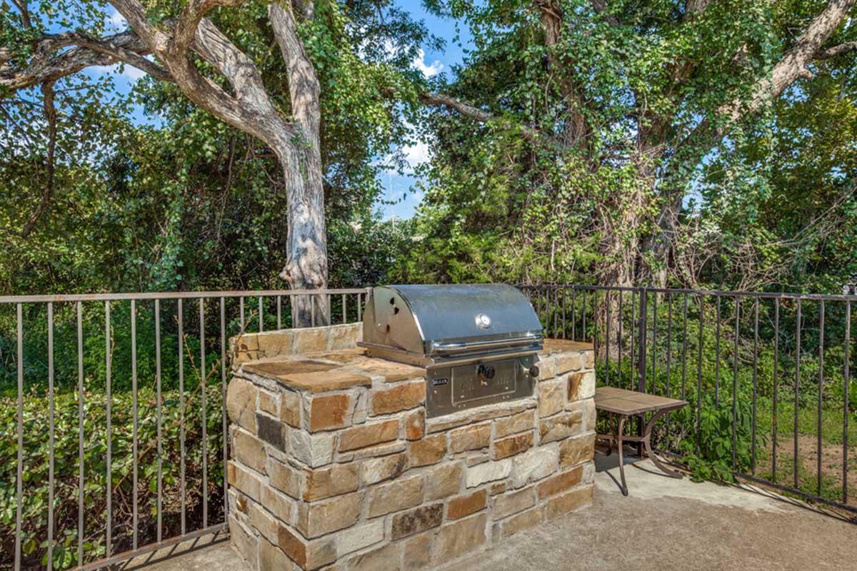 A stone-lined outdoor barbecue grill set in a natural setting, surrounded by trees. The grill is positioned on a concrete slab with a small table nearby, offering a serene environment for outdoor cooking and gatherings.