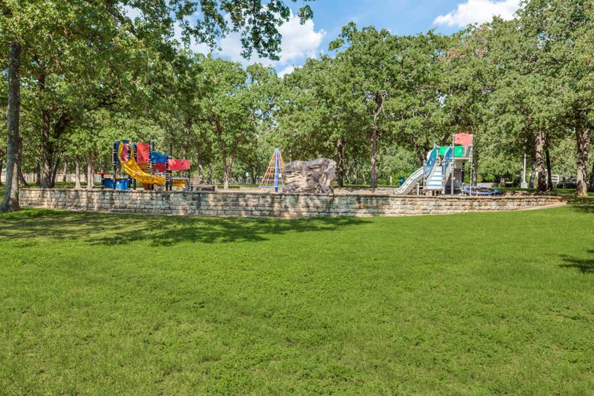 A playground area featuring colorful play structures, including slides and climbing equipment, set in a grassy area surrounded by trees under a blue sky with fluffy clouds. The space is designed for children's recreation and enjoyment.