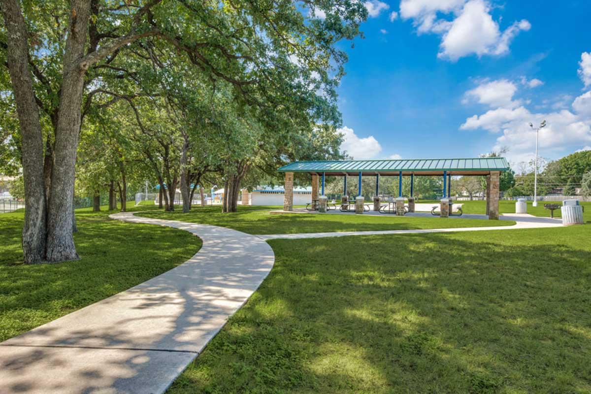 A serene park scene featuring a winding concrete pathway leading to a covered pavilion. The pavilion, with a green roof and stone walls, is surrounded by lush green grass and large trees, under a bright blue sky with fluffy white clouds. Families and friends can gather here for outdoor activities.
