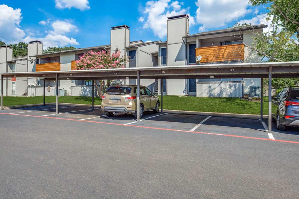 Row of apartment buildings with covered parking. Two parked cars under the carports, one tan and one blue. Lush green grass and a flowering tree in front of the buildings, with a clear blue sky and fluffy clouds above. The architecture features a mix of gray and wood accents.