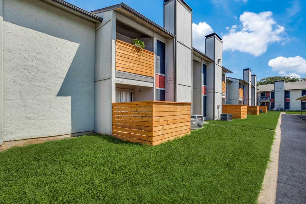 Exterior view of a multi-unit apartment building featuring wood-paneled balconies. The building has a mix of white and colored facades, with green grass in the foreground. Clear blue sky and white clouds are visible in the background, showcasing a well-maintained residential area.