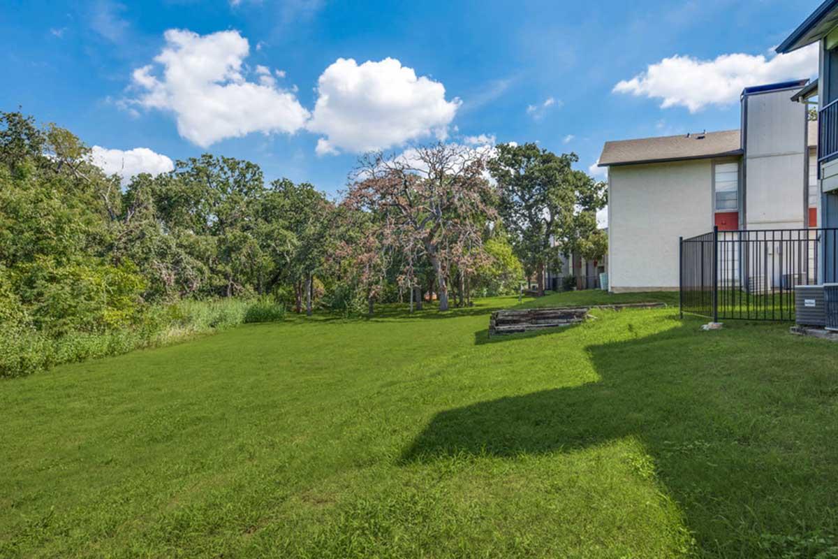 A grassy area with trees and shrubs in the background. A building is partially visible on the right side, and the sky is bright blue with a few fluffy clouds. The landscape appears well-maintained and inviting, providing a tranquil outdoor space.
