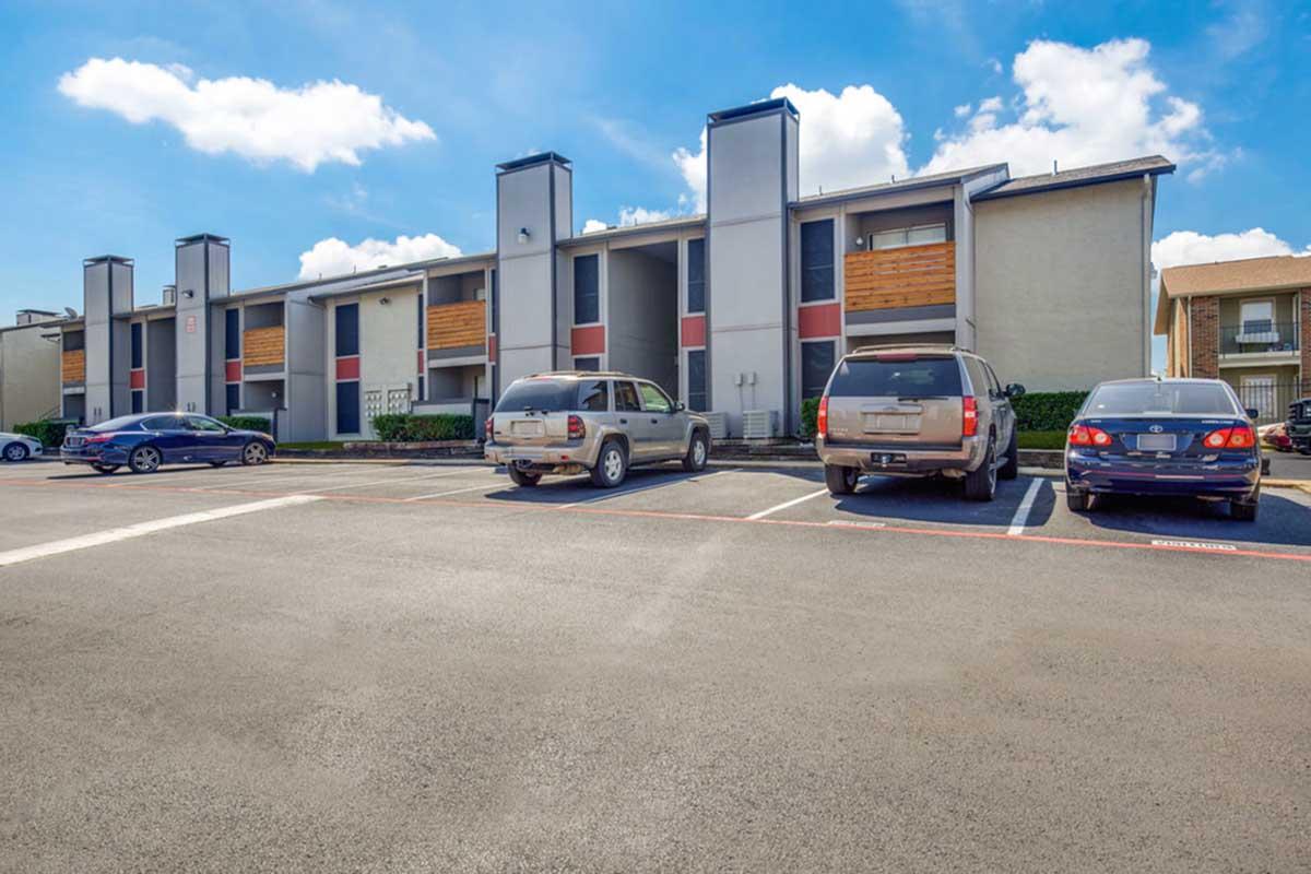 A view of a residential apartment complex featuring two-story buildings with a light exterior and wooden accents. Several parked cars are visible in the foreground, with blue skies and fluffy clouds in the background. The parking lot is neatly arranged, providing a clean and orderly appearance.