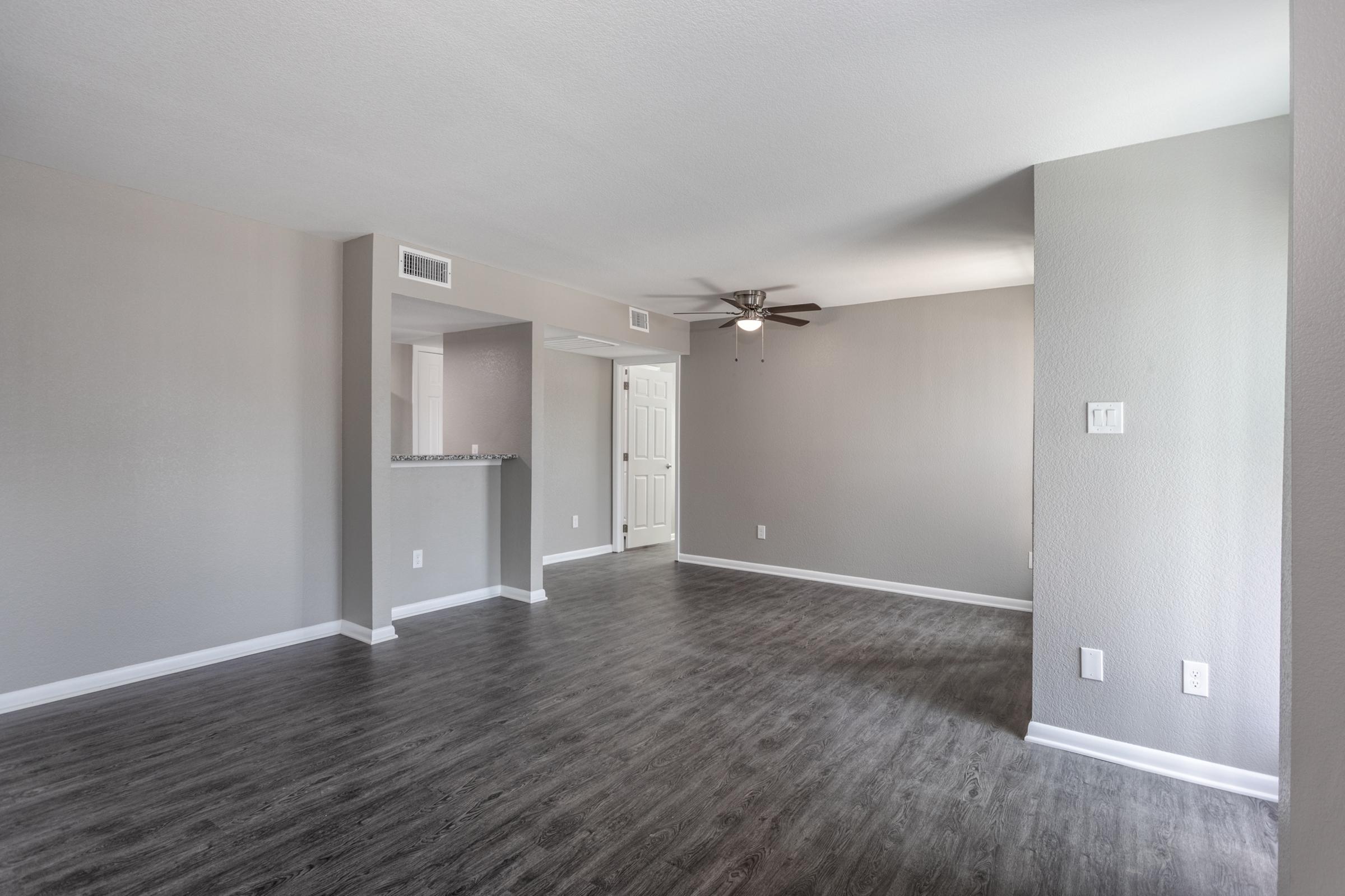 A spacious, empty living area with light grey walls and a ceiling fan. The flooring is dark wood laminate, and there is a small bar area visible leading into another room. Natural light illuminates the space, creating a bright and inviting atmosphere.