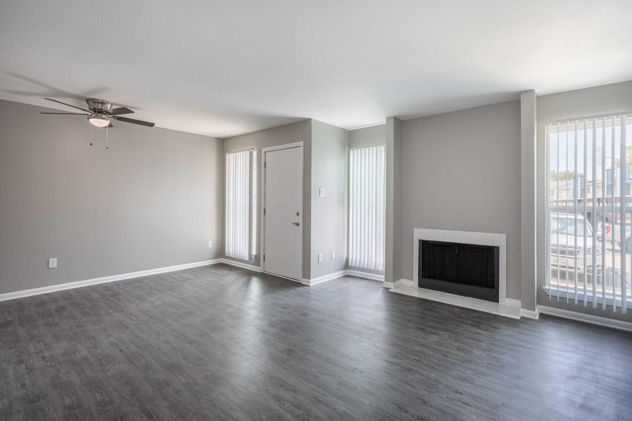 Spacious living room with light gray walls, a ceiling fan, and large windows with blinds. A white fireplace is located on one wall, and there is a front door on another wall. The flooring is dark laminate, creating a modern and inviting atmosphere.