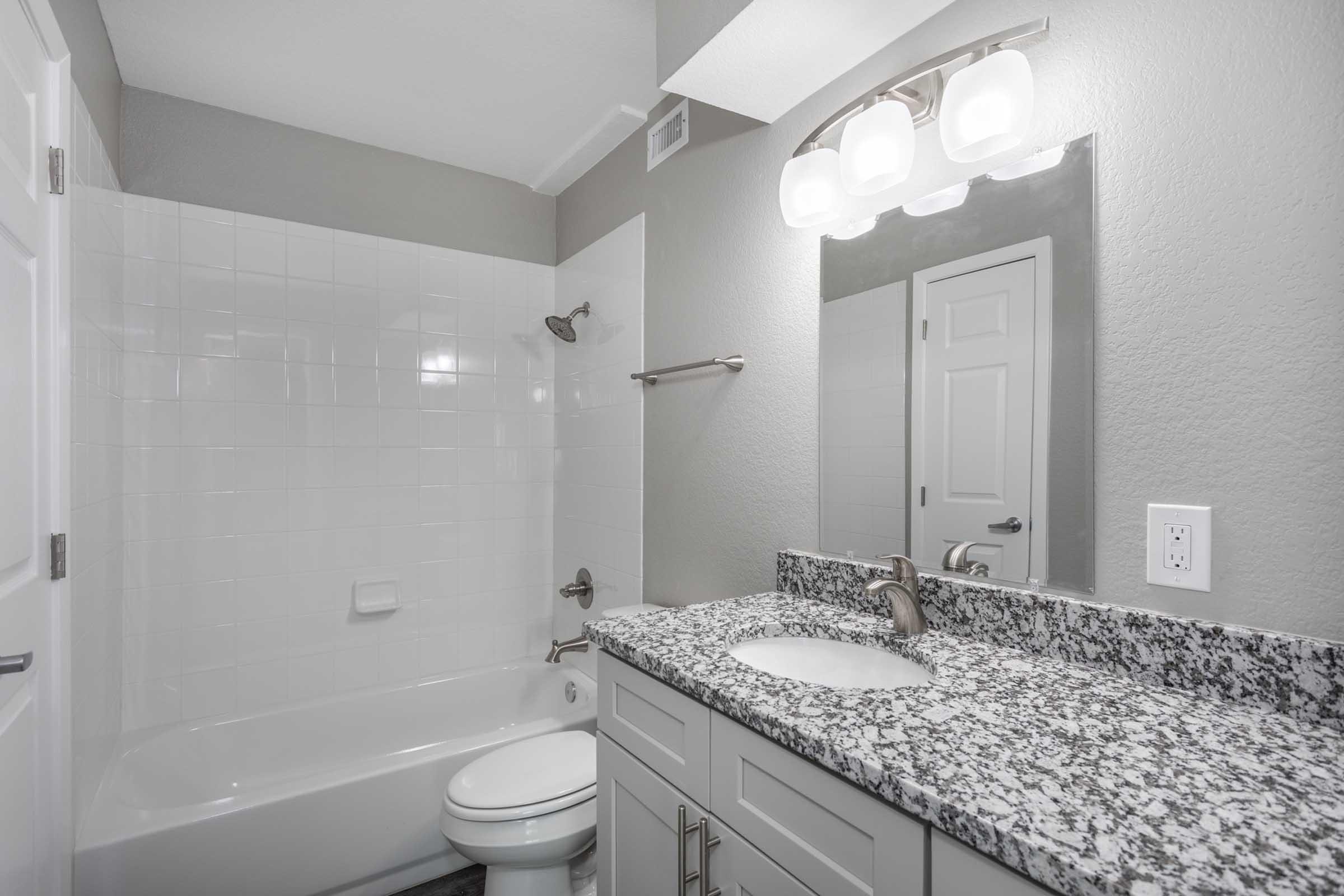 A modern bathroom featuring a white tiled bathtub and shower, a sleek granite countertop with a sink, and a large mirror above the faucet. The walls are painted in a soft gray, and there is a light fixture with three globe lights above the mirror. A towel rack is mounted next to the shower.