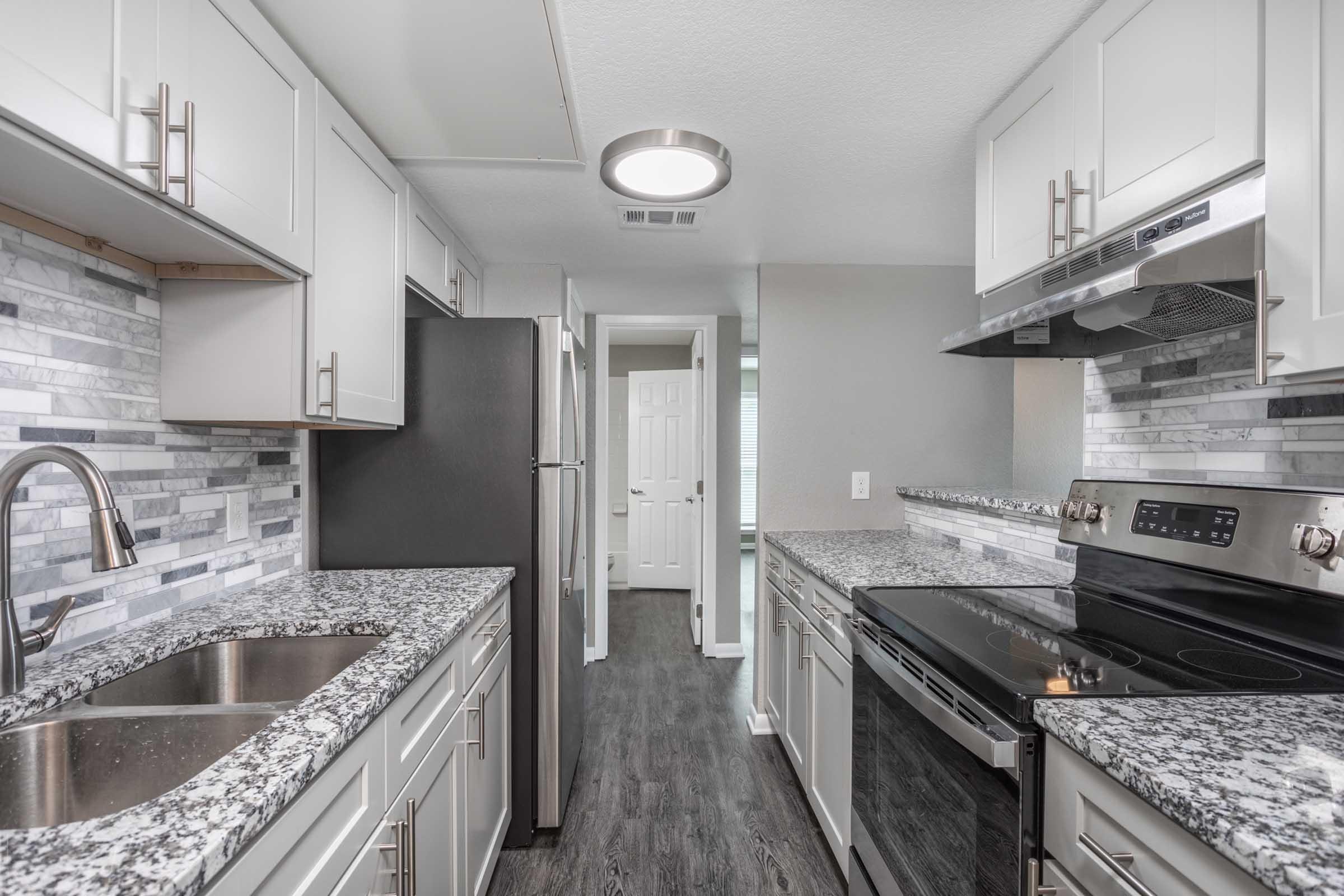 Modern kitchen featuring gray cabinetry, marble backsplash, and granite countertops. Stainless steel appliances include a refrigerator and a stove. The space has a contemporary design with a sleek look and ample natural light, creating a bright and inviting atmosphere.