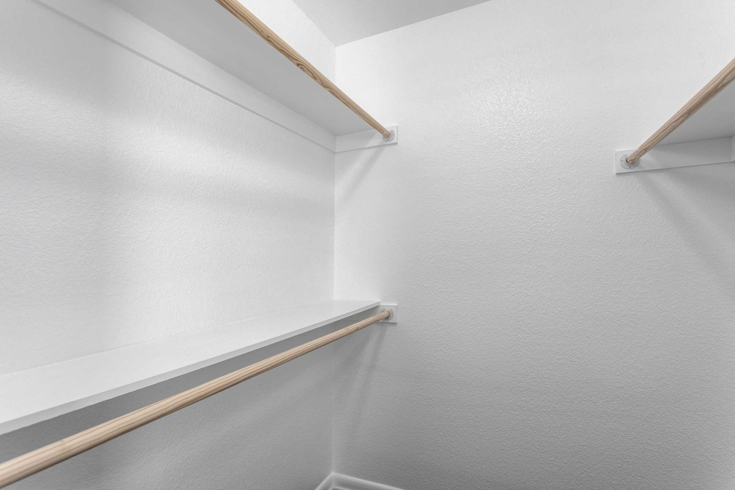 An empty closet with white walls, featuring two wooden shelves and a hanging rod on each side. The space is well-lit, highlighting the simplicity and cleanliness of the design.
