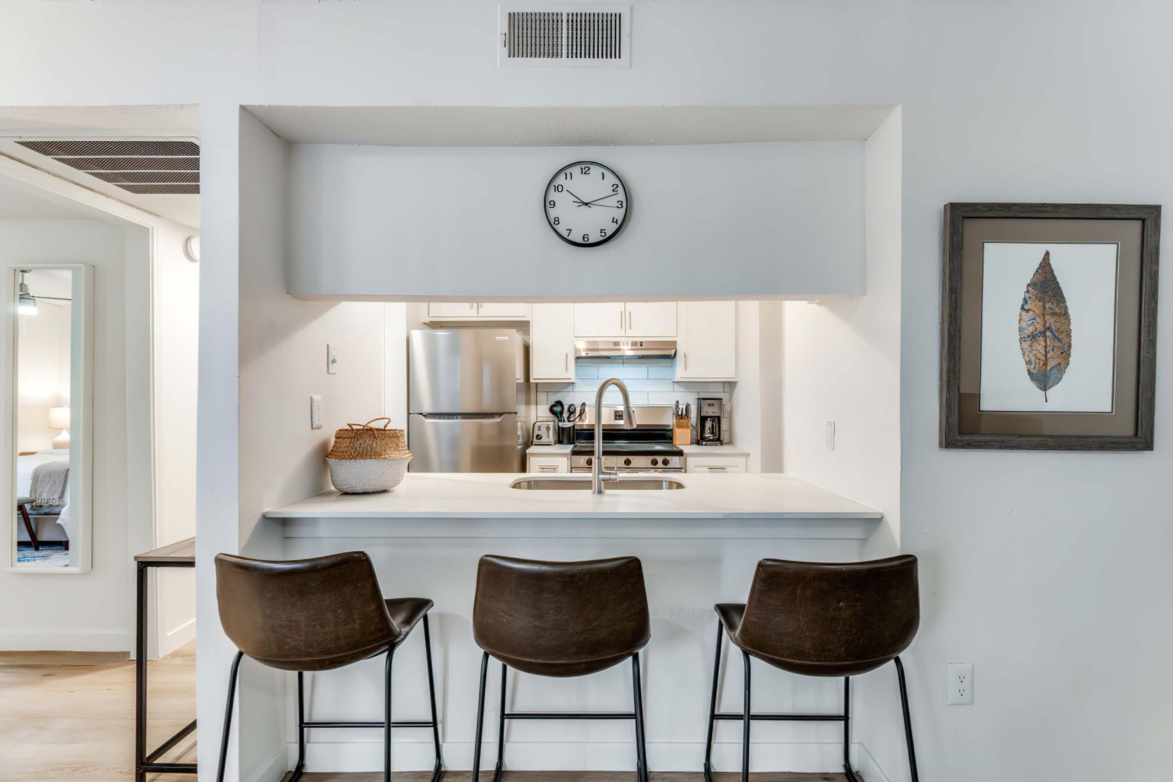 Modern kitchen with a white countertop and bar seating for three. A clock on the wall shows the time, while stainless steel appliances, including a refrigerator and sink, are visible. A framed artwork of a leaf hangs nearby, enhancing the contemporary decor.
