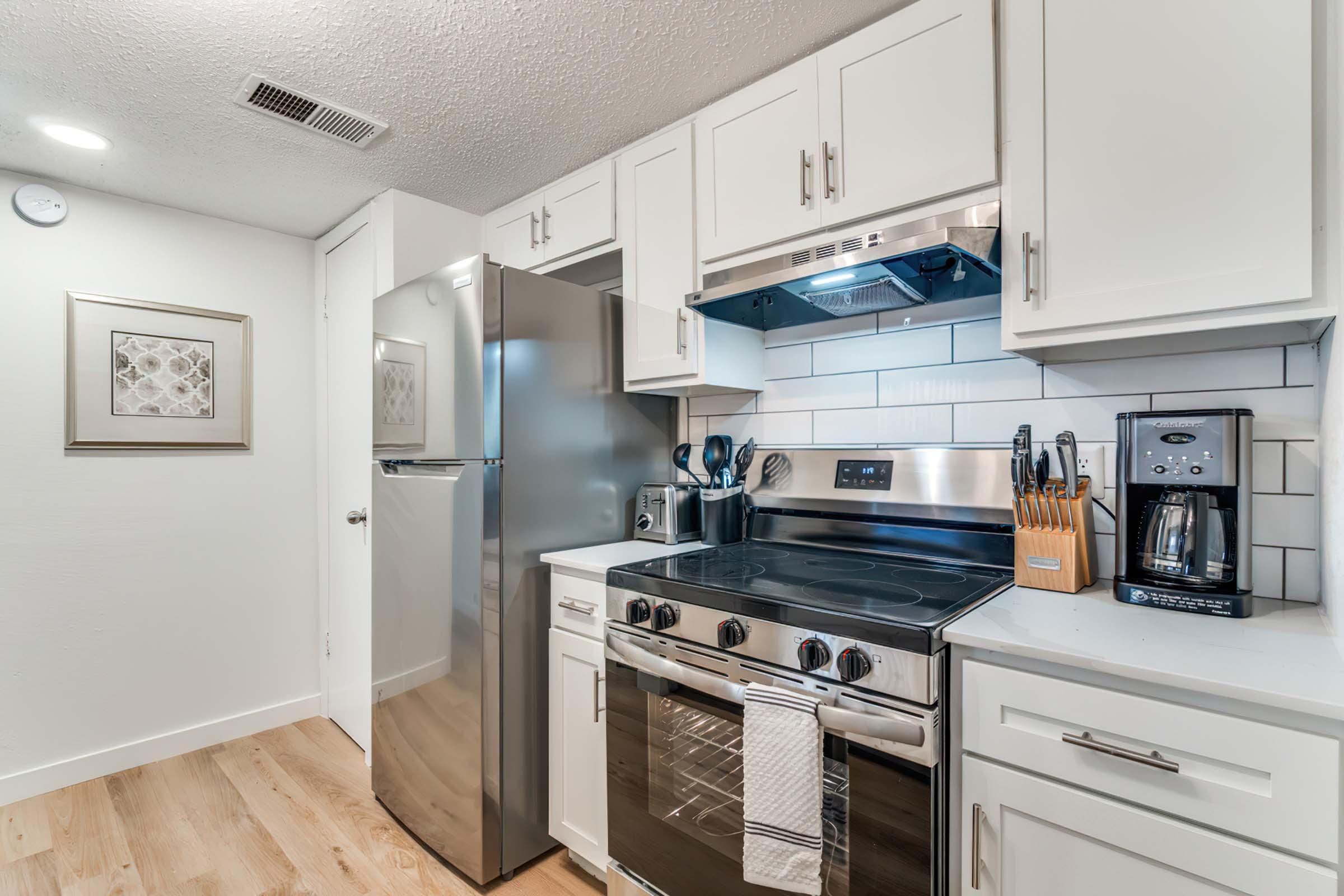 Modern kitchen featuring stainless steel appliances, including a refrigerator and stove. The cabinetry is white with a sleek design, while the countertop is light-colored. There are kitchen utensils and a coffee maker on the counter, with a tiled backsplash enhancing the aesthetic. The space is well-lit and tidy.