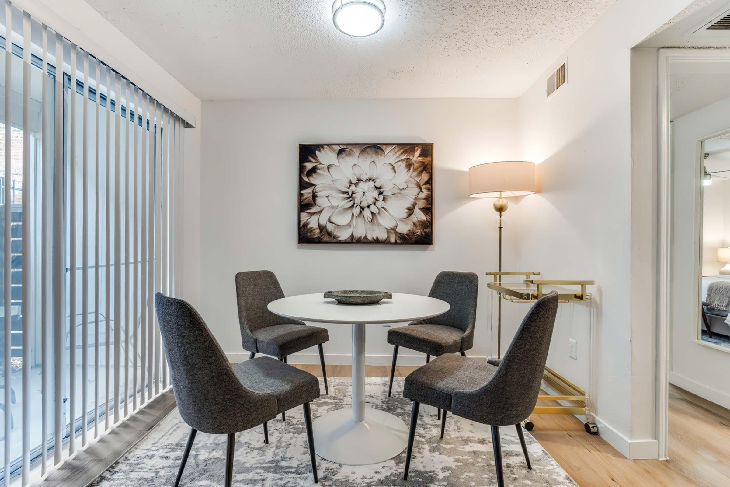 A modern dining area featuring a round white table surrounded by four upholstered gray chairs. A large floral artwork hangs on the wall, and a stylish floor lamp stands beside the table. The space has soft lighting and a light wood floor, creating a welcoming atmosphere. Large sliding glass doors lead to an outdoor area.