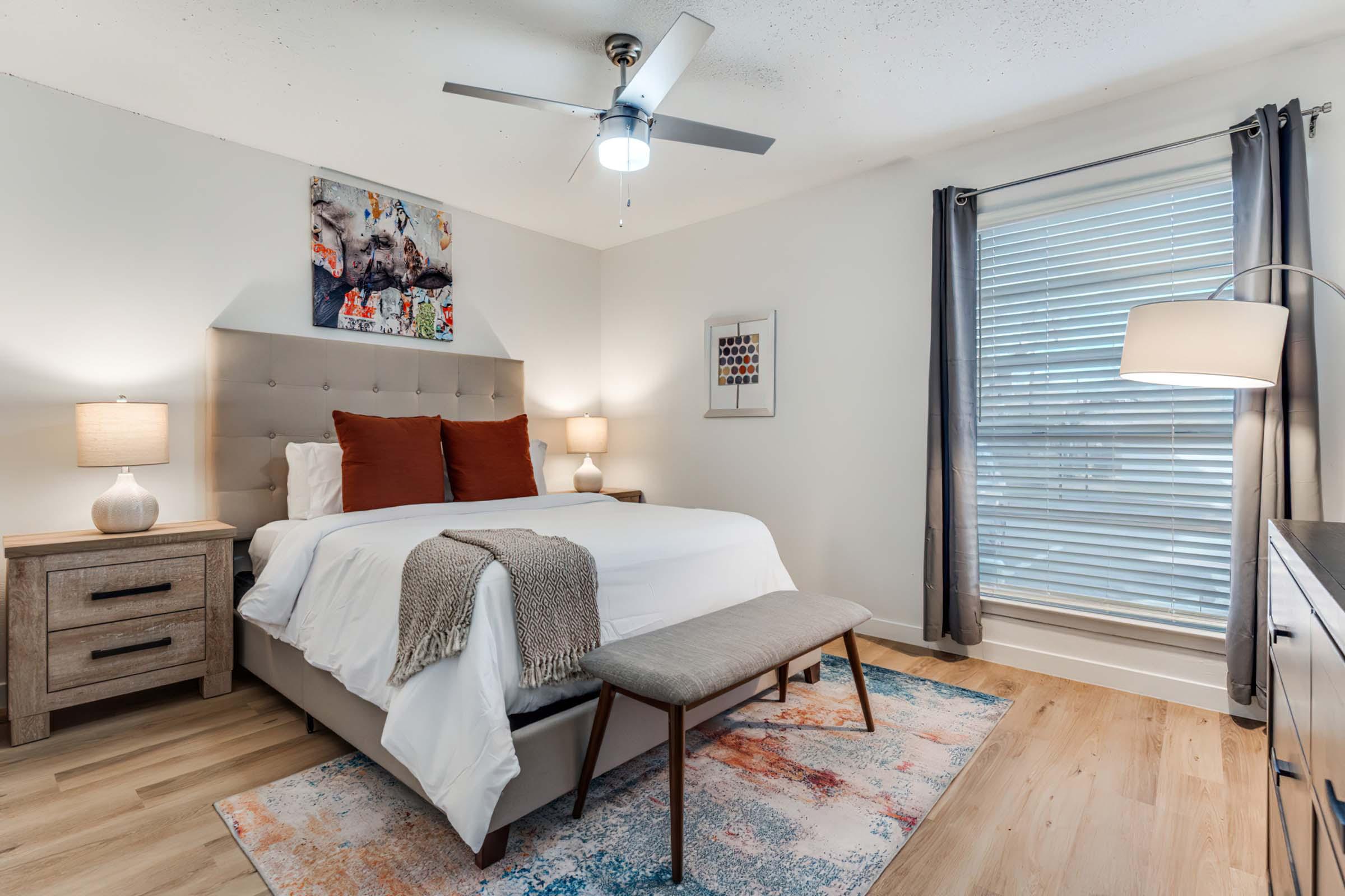 A cozy bedroom featuring a queen-sized bed with white bedding and decorative pillows, a stylish bench at the foot of the bed, a colorful area rug, bedside lamps, and a ceiling fan. Natural light streams in through a window with blinds, and there is artwork on the wall, adding a welcoming atmosphere.