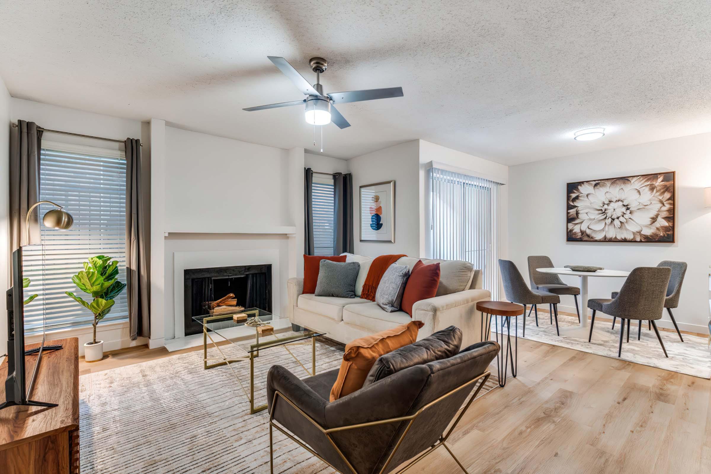 Cozy living room with a modern design featuring a white sofa with colorful throw pillows, a stylish armchair, and a coffee table. A fireplace adds warmth, while large windows with blinds allow natural light. A dining area with a round table and chairs is visible in the background, enhancing the inviting atmosphere.