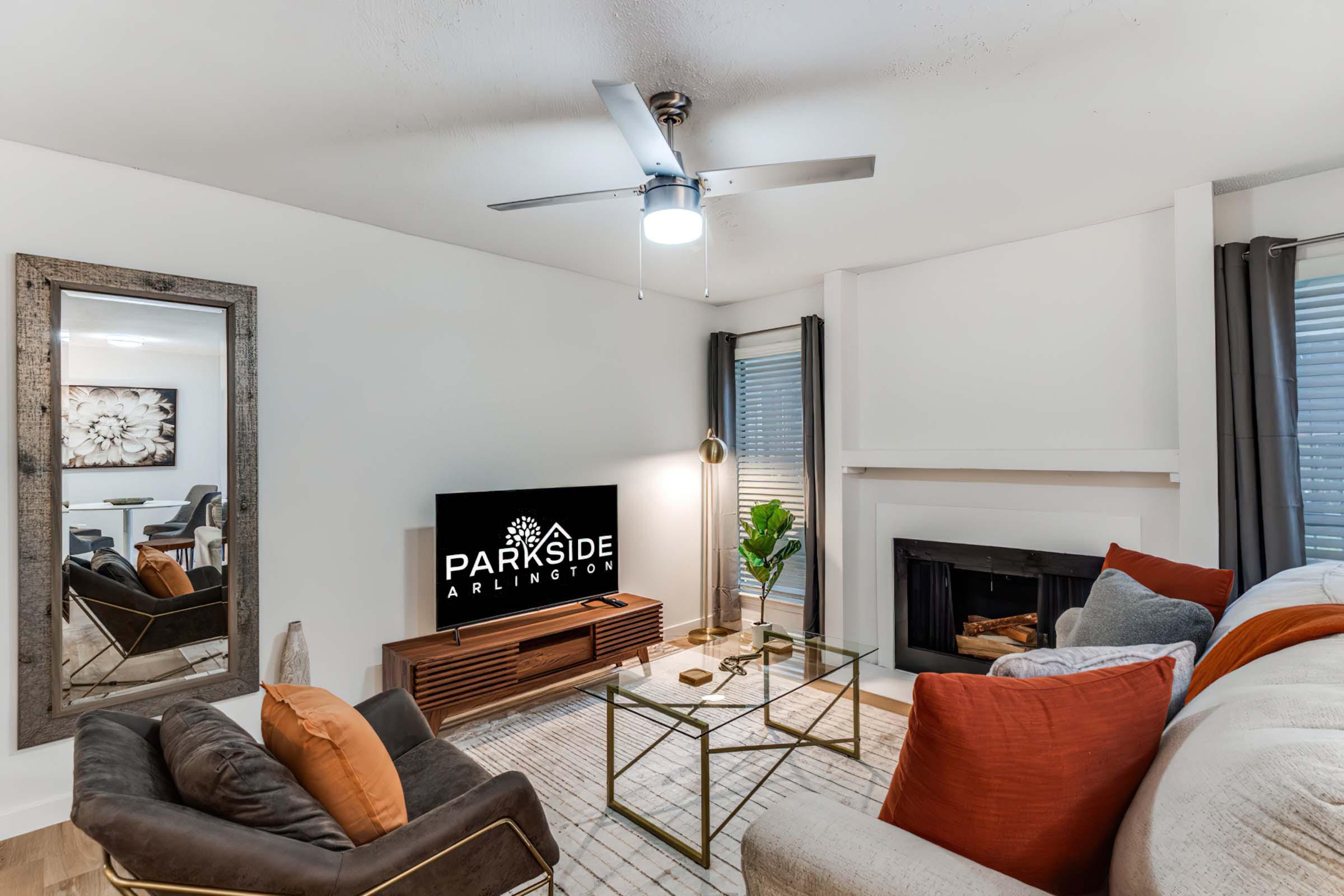 A modern living room featuring a comfortable couch with colorful pillows, a glass coffee table, a sleek TV stand with a television displaying the "PARKSIDE ARLINGTON" logo, and a stylish ceiling fan. There are decorative plants and artwork on the walls, creating a cozy atmosphere.