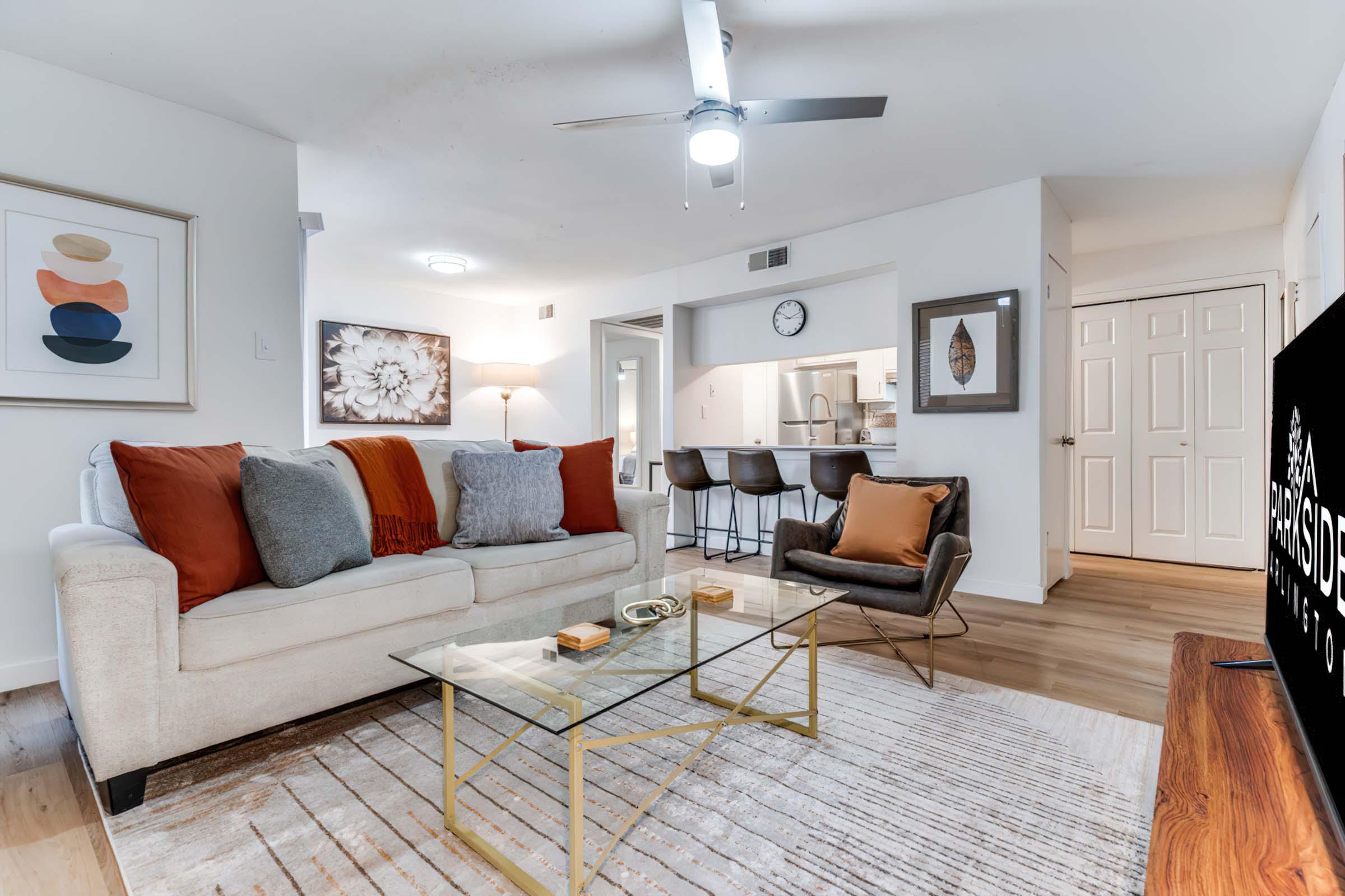 A cozy living room featuring a beige sofa adorned with orange and gray pillows, a glass coffee table, and a small accent chair. There's a ceiling fan overhead and framed artwork on the walls. A TV stand with a television is visible, and the open layout leads to a kitchen area in the background.