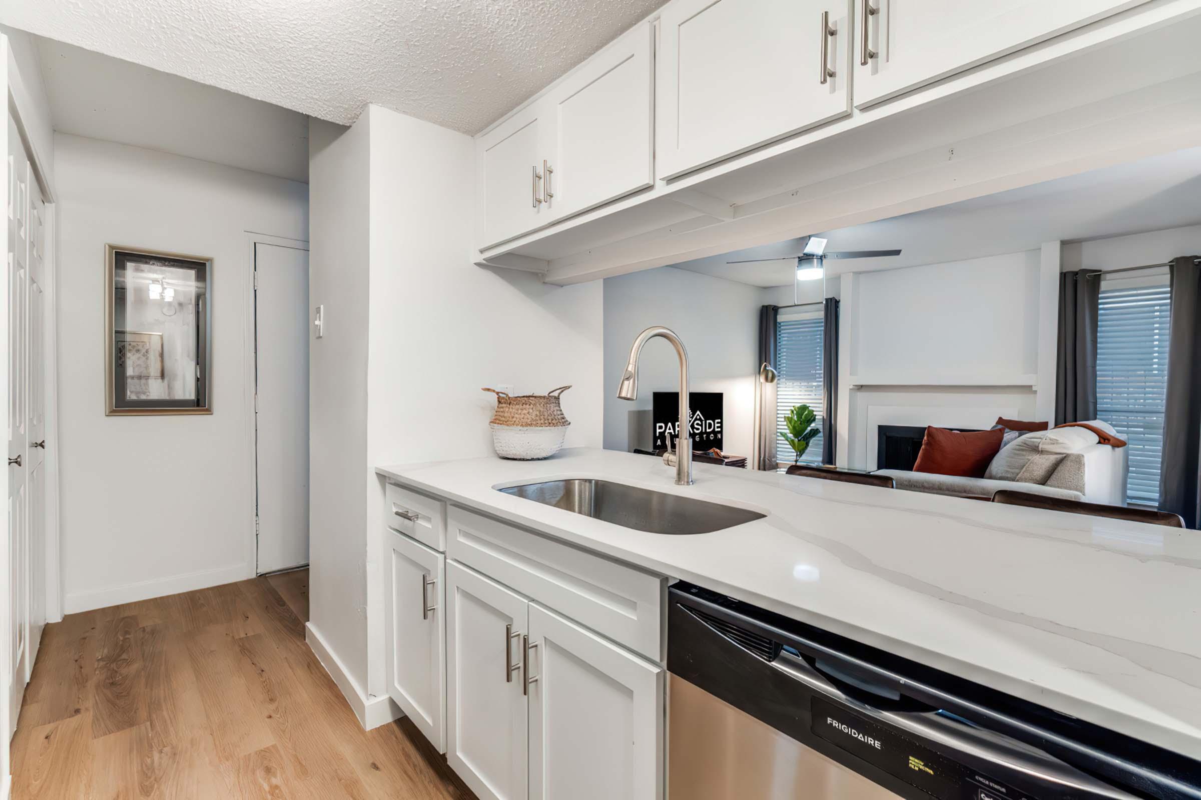 A modern kitchen with white cabinetry, featuring a stainless steel sink and dishwasher. The open space leads to a cozy living area with a fireplace, decorated with pillows and plants. Bright natural light streams in through windows, creating a welcoming atmosphere. Light wood flooring throughout.