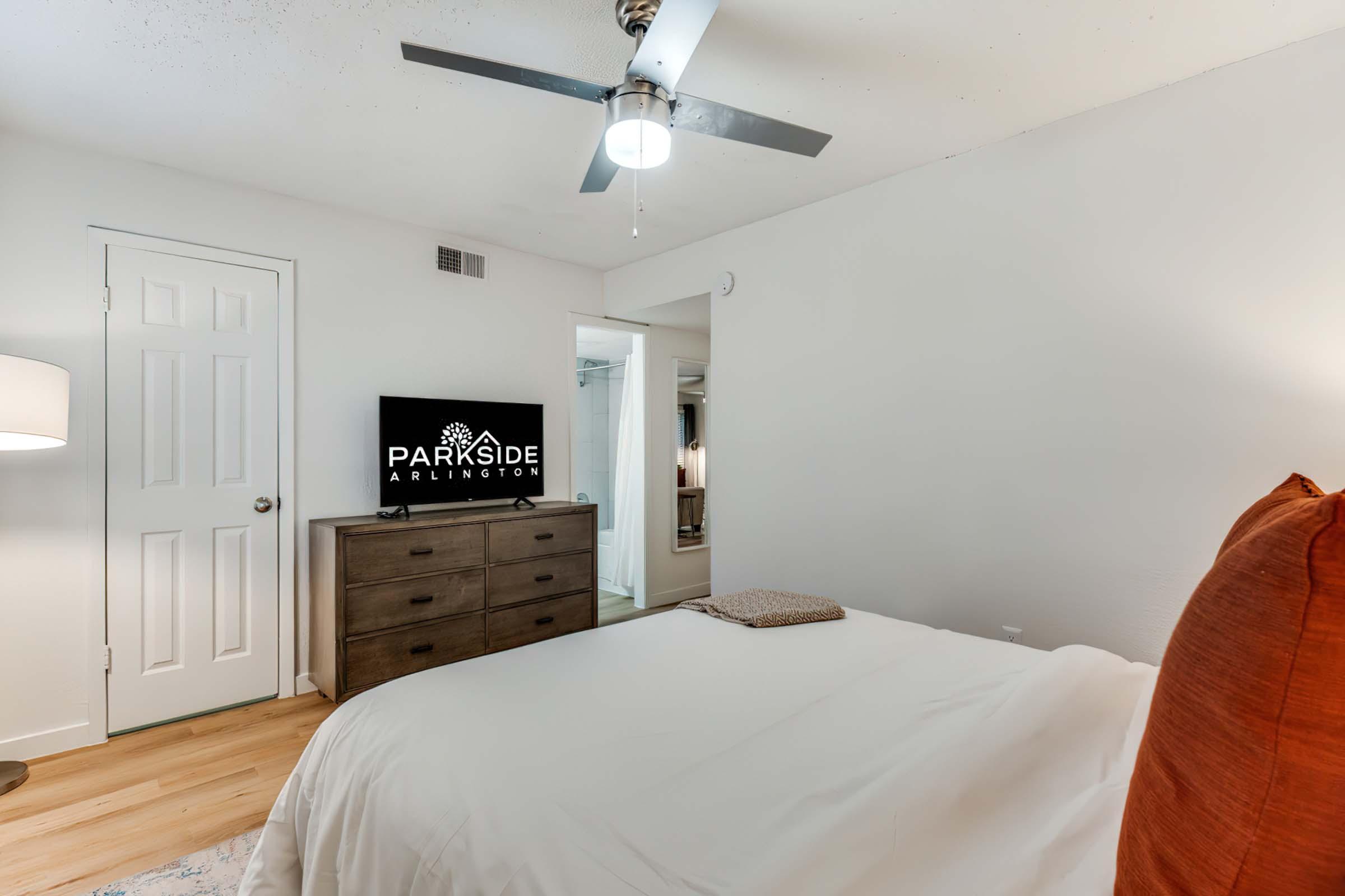 A modern bedroom featuring a neatly made bed with white bedding, a wooden dresser with a television on top, a ceiling fan, and a lamp. A closet door is visible on the left, and there’s a large mirror in the background reflecting a well-lit, minimalist space.