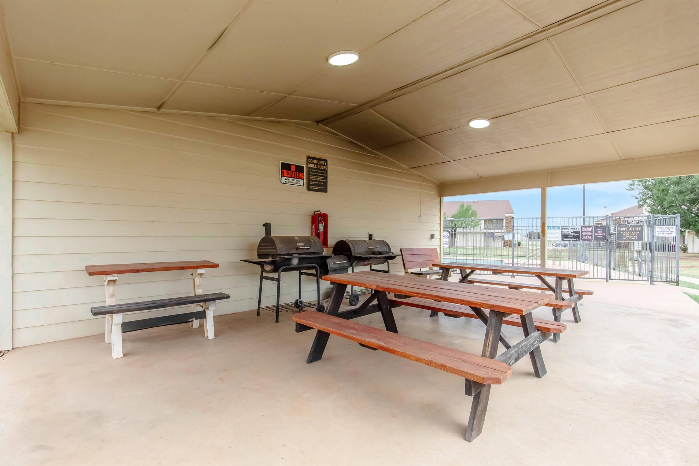 Covered outdoor area featuring several picnic tables made of wood, with two charcoal grills on one side. There is a fire extinguisher mounted on the wall. The space is well-lit and offers a view of a fence and grassy area outside.