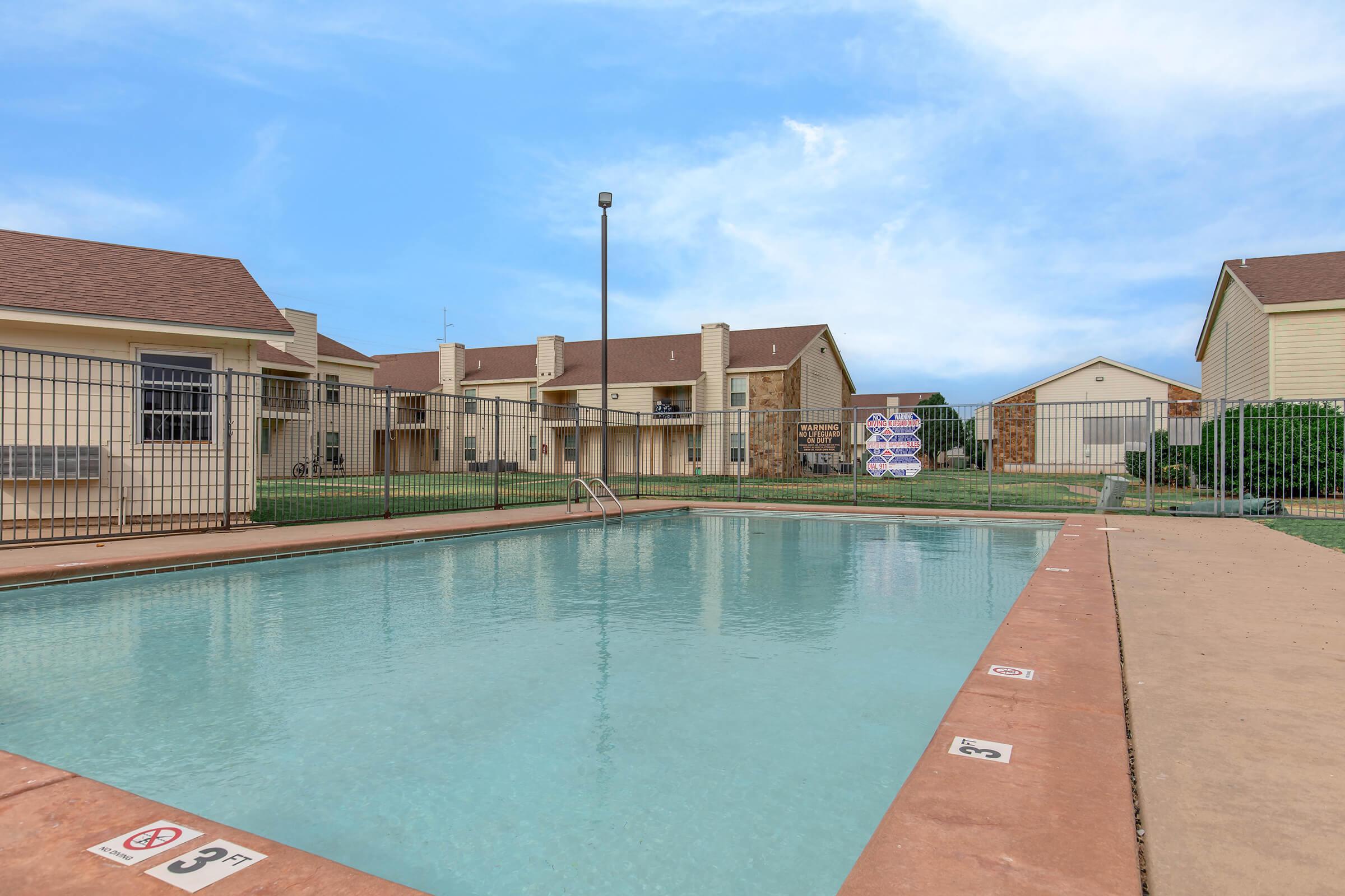 A clean, inviting swimming pool surrounded by a fence, with lounge areas nearby. Several apartment buildings are visible in the background under a clear blue sky. The pool deck is marked with depth indicators and features a light pole at one end. Green grass and shrubs border the area, creating a pleasant outdoor space.