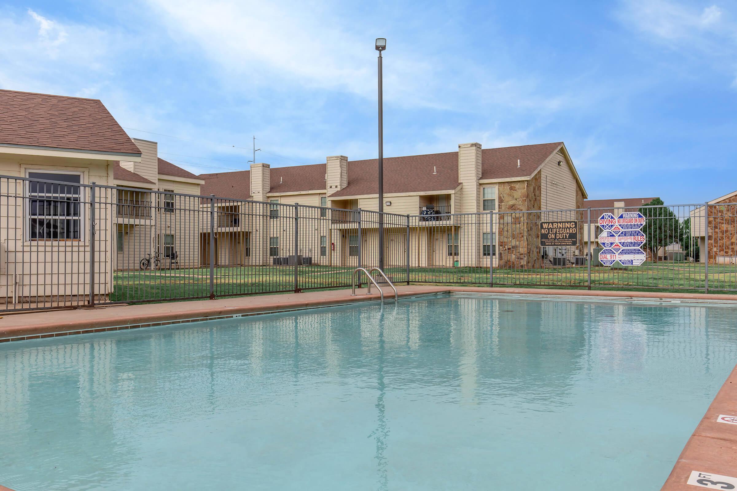 A clear swimming pool with ladders, surrounded by a gated fence. In the background, there are residential buildings with balconies and brown roofs. The sky is bright with some clouds, and there are warning signs visible near the pool area. The grass is well-maintained.
