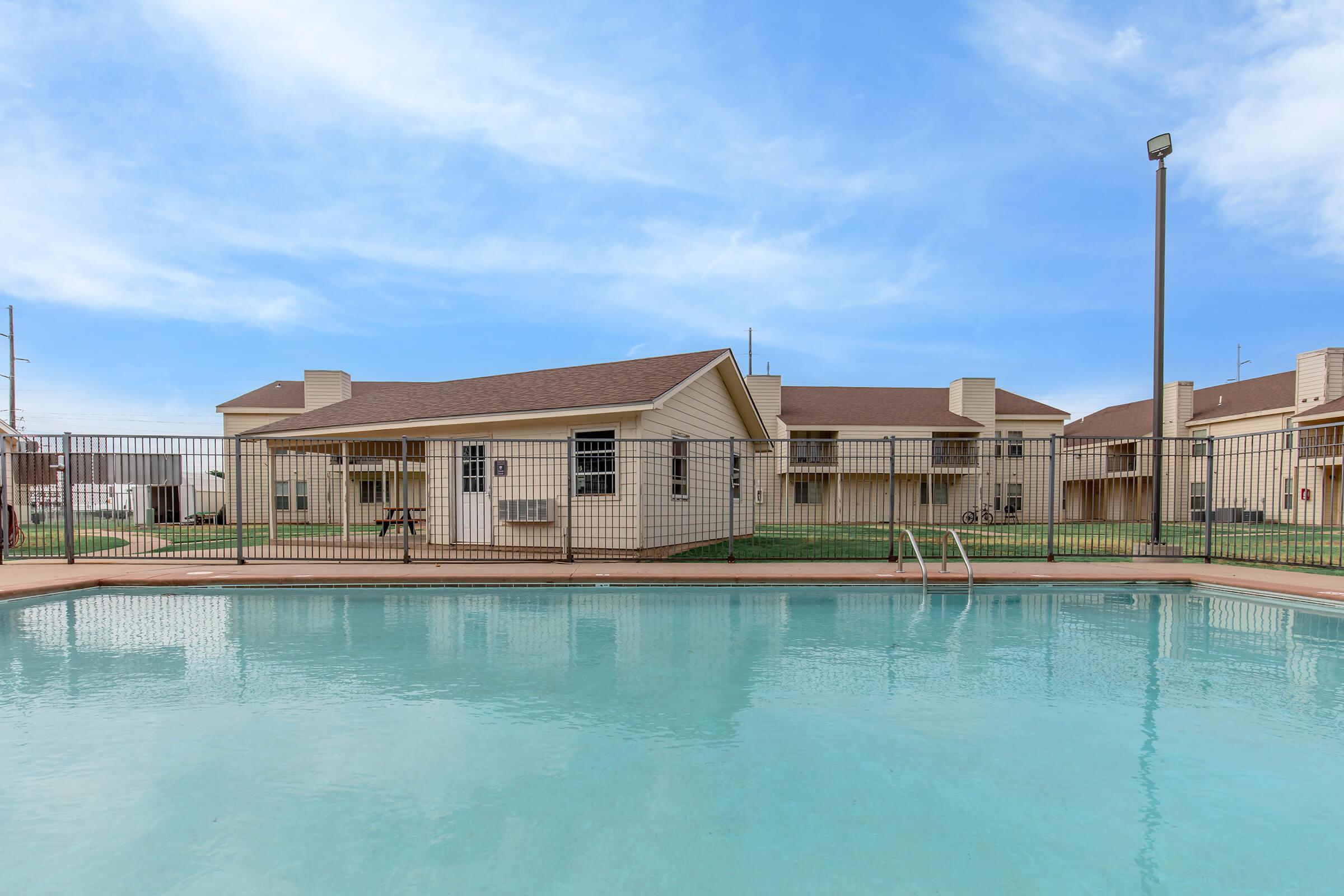 A clear swimming pool surrounded by a fenced area, with beige buildings in the background under a blue sky. The pool has a ladder for entry, and the grass around the pool area is well-maintained. The scene gives a serene and inviting atmosphere.