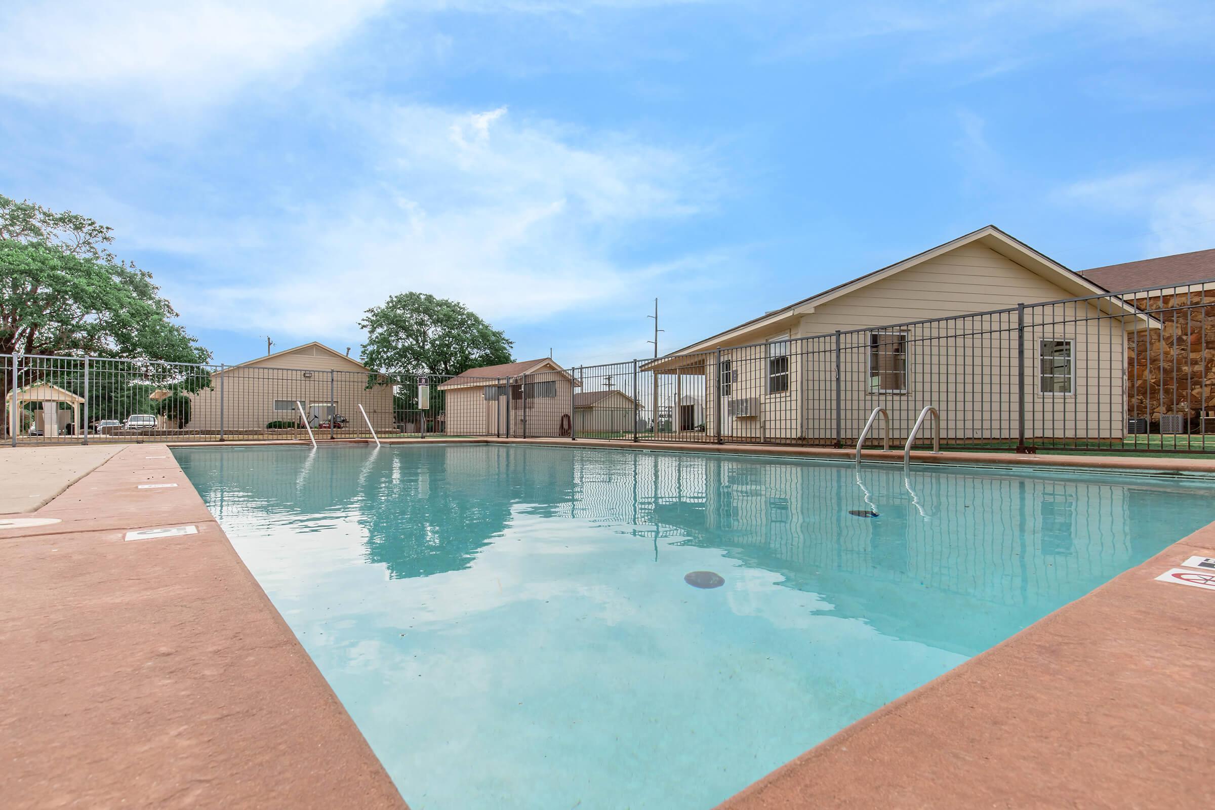 A clear swimming pool surrounded by a gated area, with several residential buildings in the background. The sky is bright with a few clouds, and there are trees nearby, creating a serene outdoor atmosphere. The pool deck is made of beige concrete with pool ladders visible.