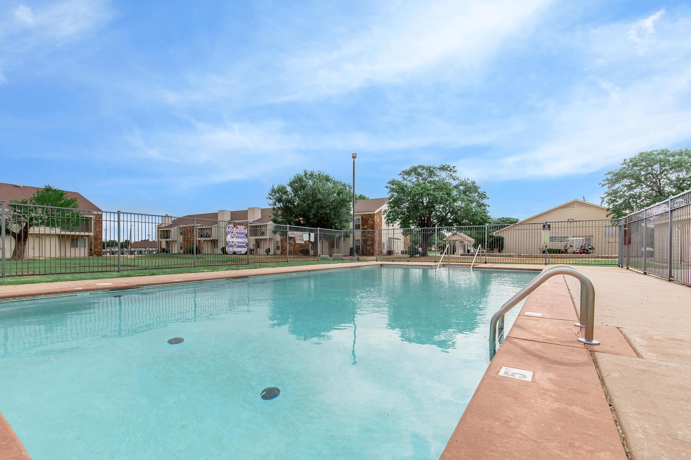 A clear blue swimming pool with a diving board, surrounded by a fence and grassy area. In the background, there are a few residential buildings and trees, under a bright blue sky with some clouds. The pool area appears well-maintained and inviting.