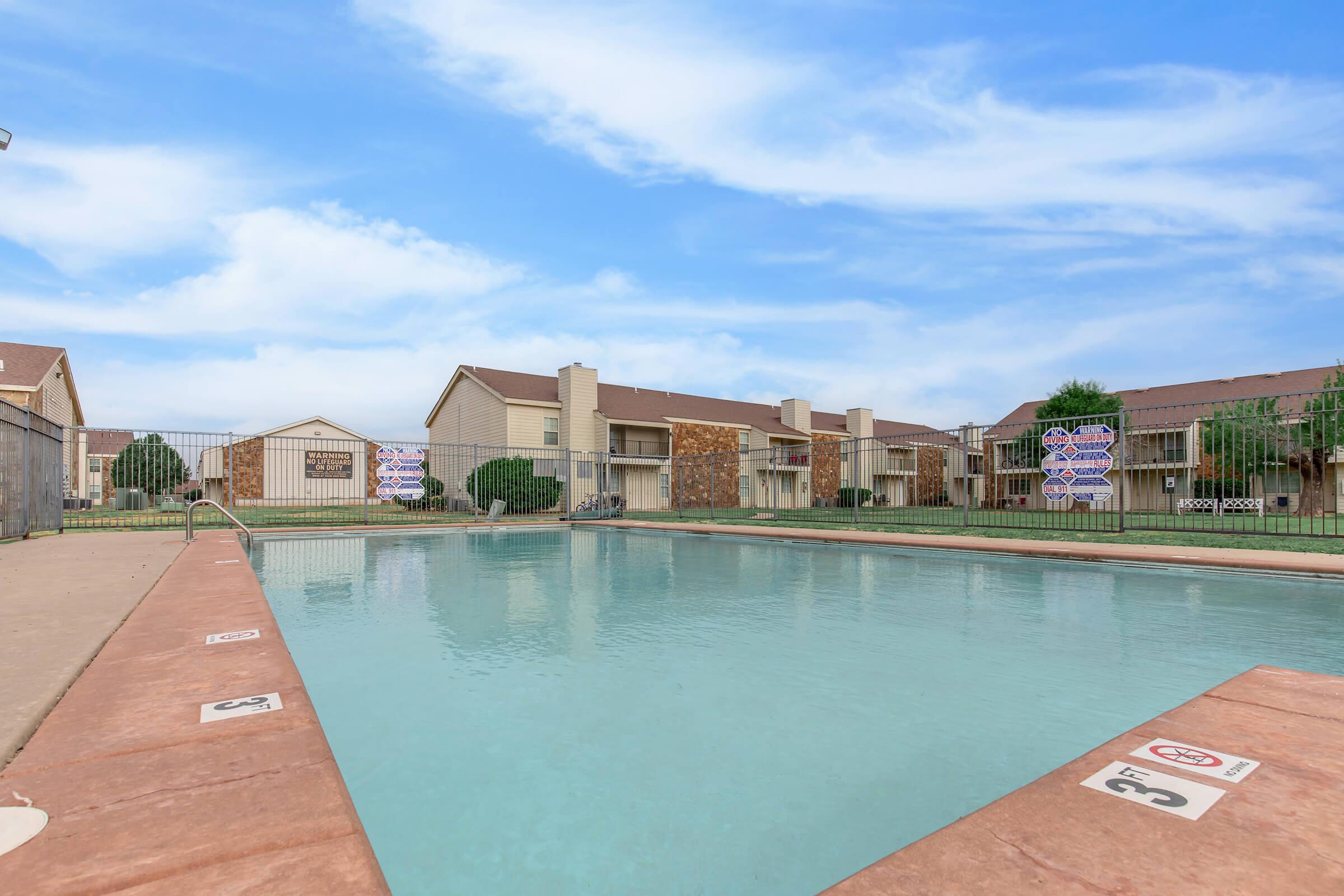 A clear swimming pool surrounded by a fenced area, with several apartment buildings in the background. The sky is blue with some clouds, and there are signs posted around the pool area. Lush greenery can be seen along the edges of the pool.