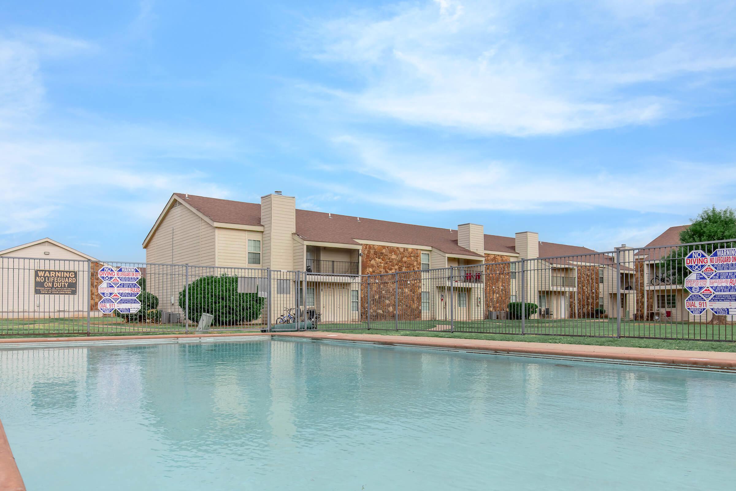 A view of an apartment complex with a swimming pool in the foreground. The pool is enclosed by a fence, and there are warning signs posted. The buildings feature a mix of stone and siding, with clear blue skies in the background.
