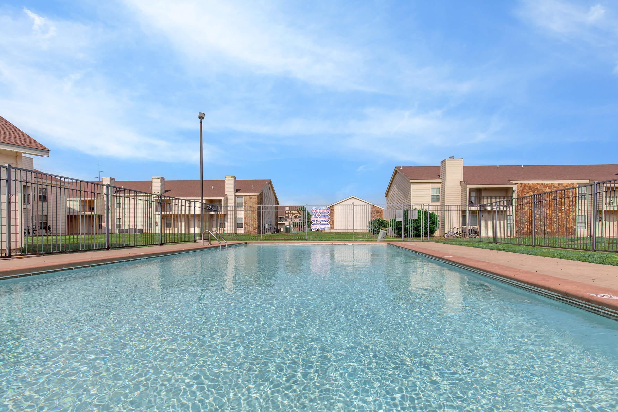 A clear swimming pool surrounded by a grassy area and two-story apartment buildings. The sky is blue with a few clouds, and the pool area is enclosed by a fence. The scene conveys a tranquil atmosphere ideal for relaxation.