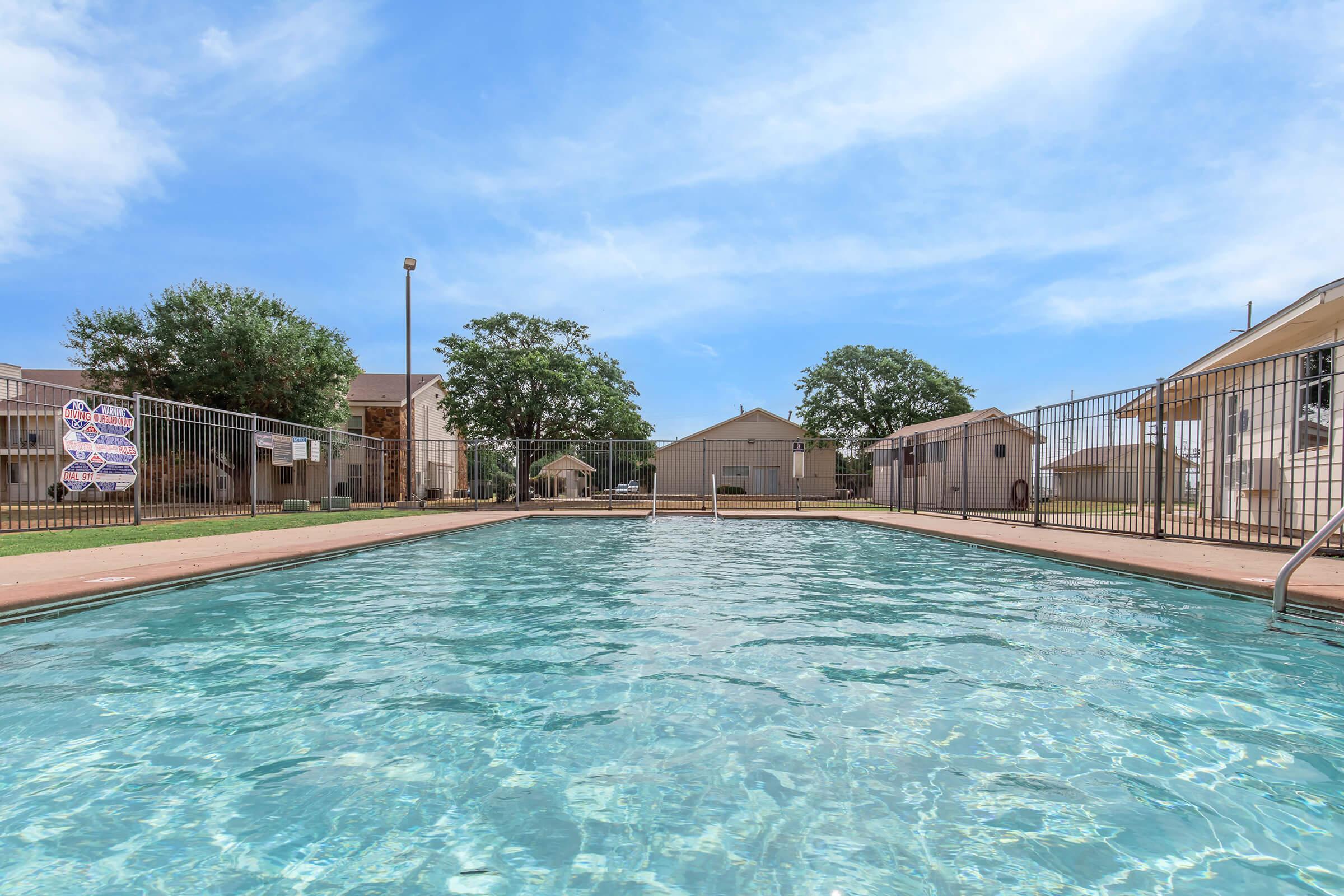 A clear swimming pool surrounded by a fenced area, with lush green grass and trees in the background. The pool is filled with sparkling water, reflecting the blue sky. Residential buildings can be seen on either side of the pool, indicating a suburban setting.
