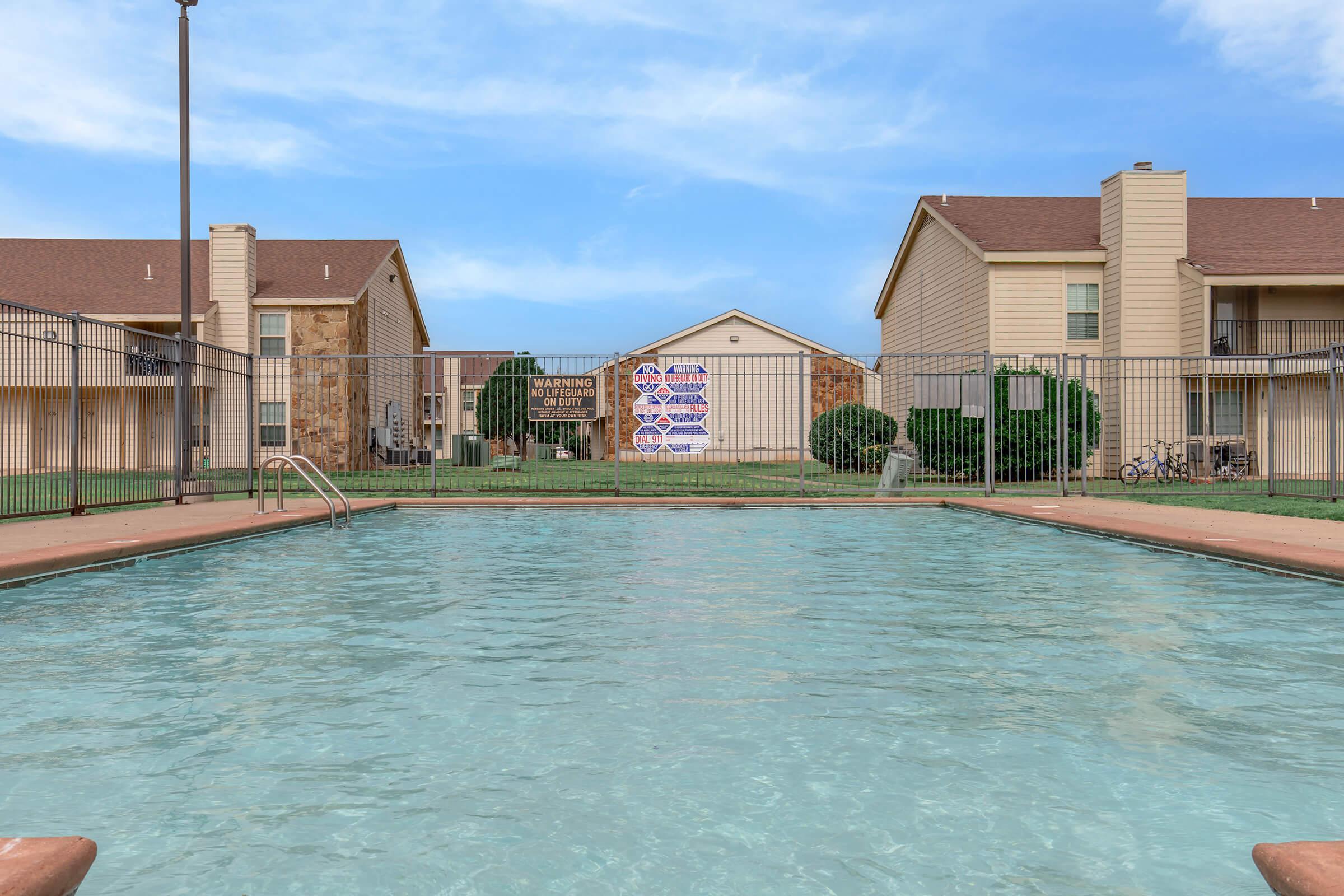A view of a swimming pool in an apartment complex, surrounded by a fenced area. Two buildings flank the pool, with a grassy lawn and bicycles visible in the background. Signs are posted on a building in the distance, indicating community rules or announcements. The sky is clear and blue.