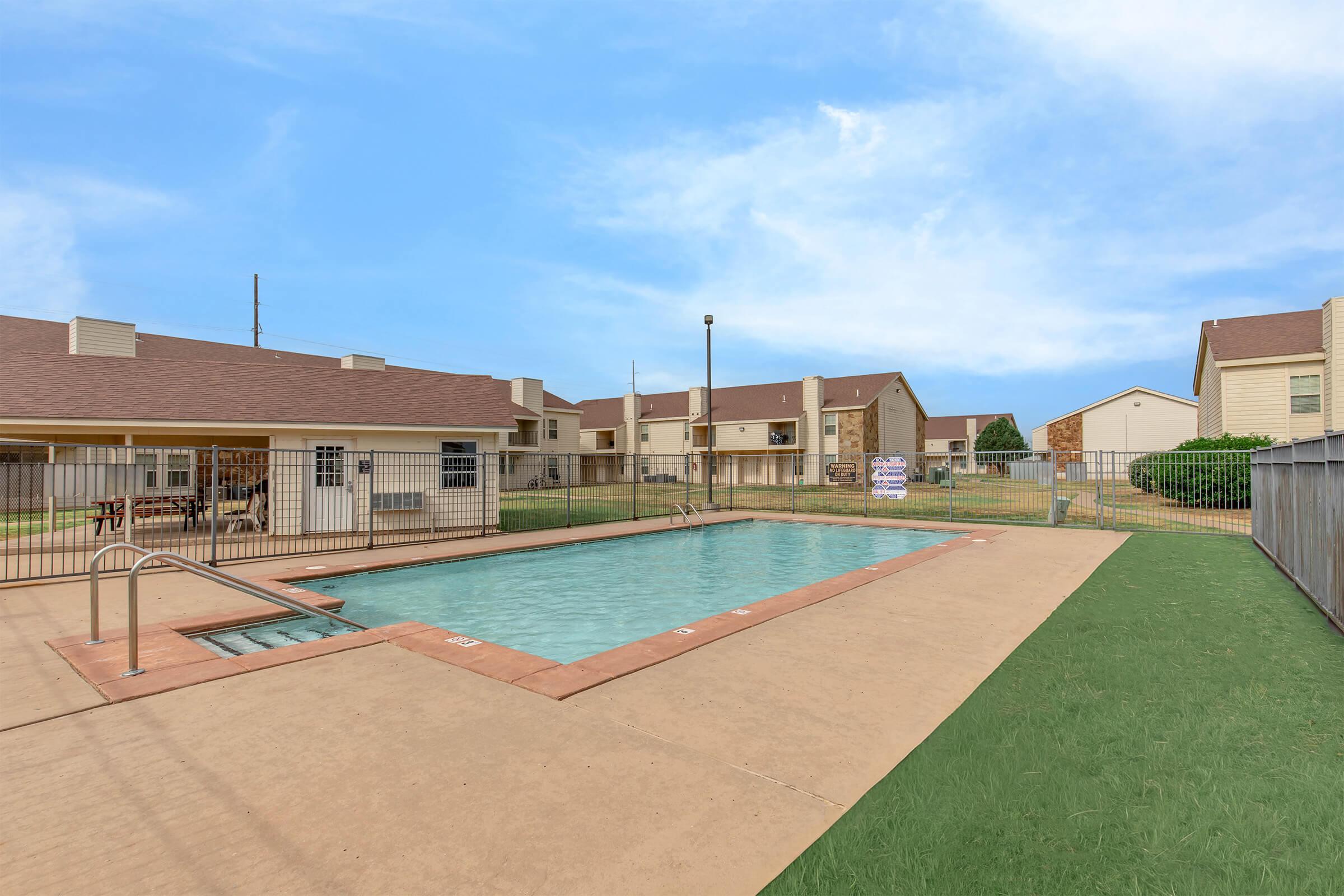 A clear swimming pool surrounded by a fenced area, set in a residential complex. There are several buildings with brown roofs in the background, and a well-maintained lawn with green grass around the pool area. The sky is blue with a few clouds, indicating a pleasant day.