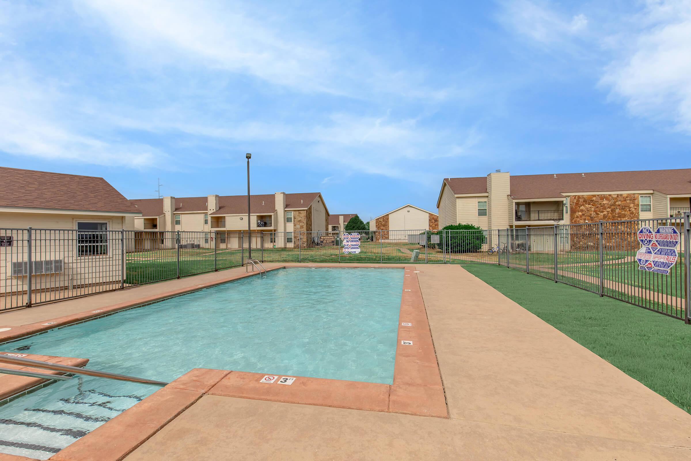 A clean outdoor swimming pool surrounded by a fence, with well-maintained landscaping. In the background, there are several residential buildings with a blue sky overhead. The pool area features lounge space and is not currently occupied.