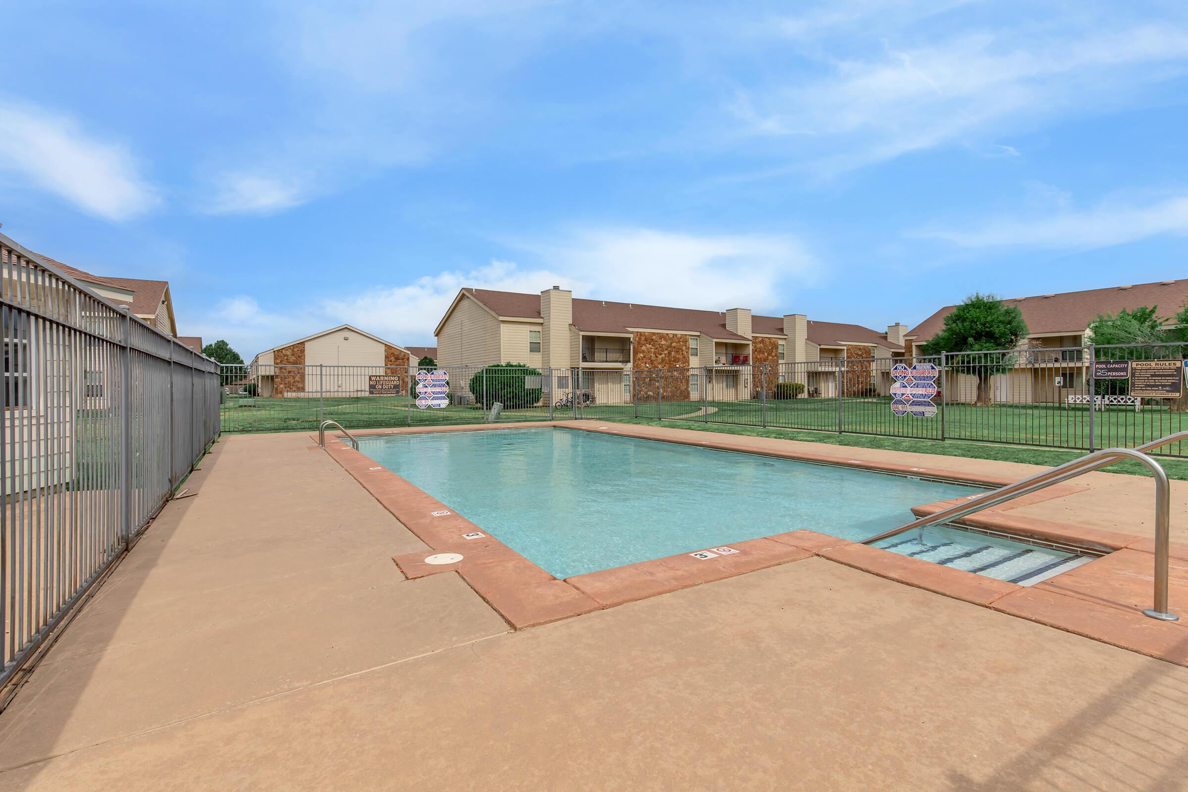 A clear swimming pool surrounded by a fence, set in a green area with apartment buildings in the background. The sky is bright and partly cloudy, and there are signs posted nearby. The pool features a staircase for entry and has a clean and inviting appearance.