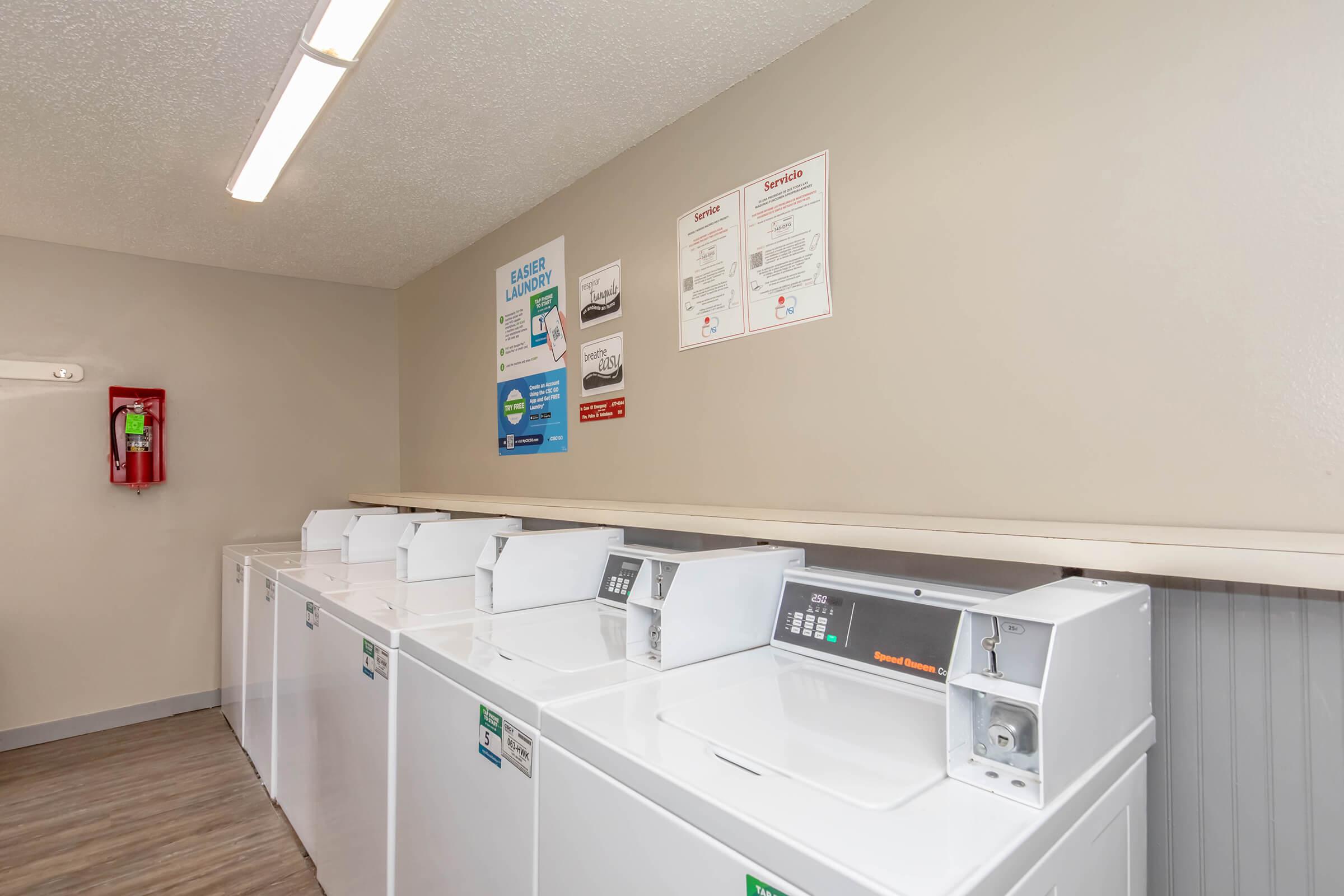 A well-lit laundry room featuring several white washing machines and dryers aligned against a beige wall. Posters on the wall provide laundry instructions and safety information. A fire extinguisher is mounted nearby for safety. The floor is made of light wooden planks.