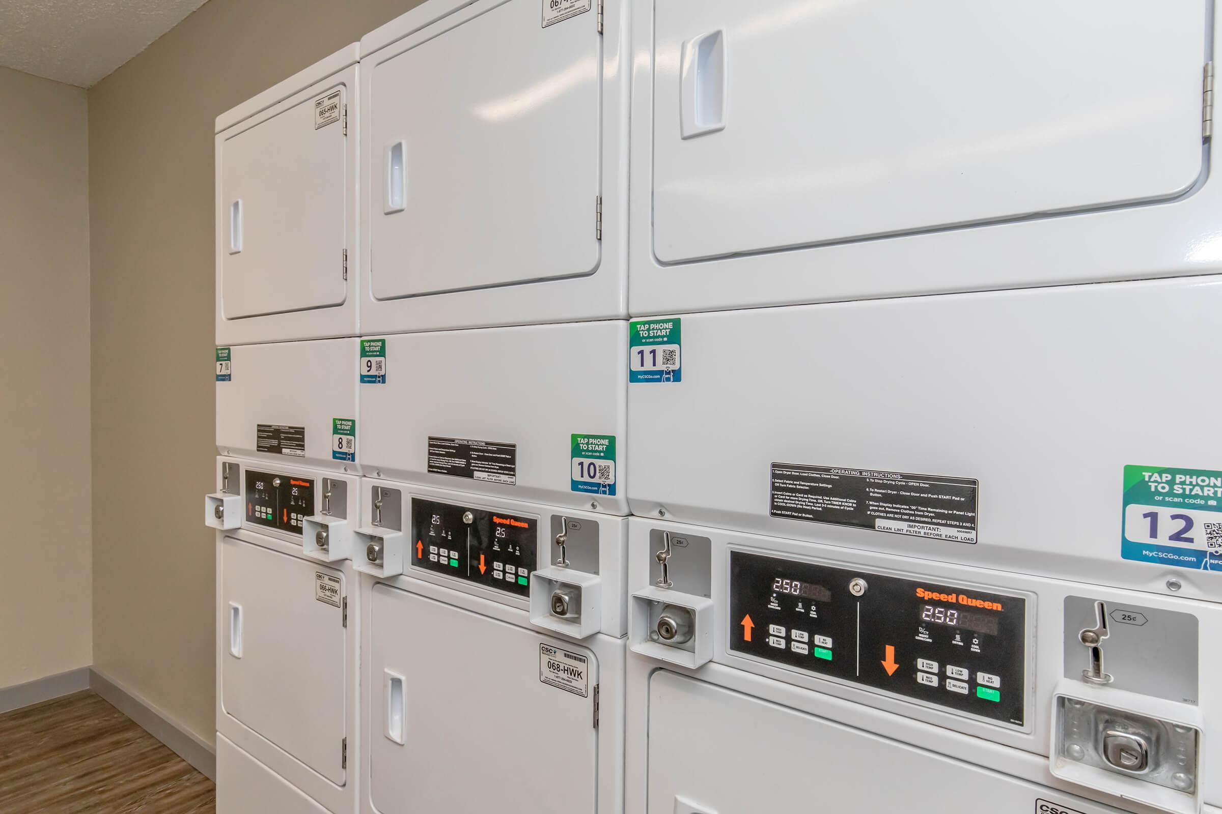 A row of white laundry machines with digital displays and buttons for operation, located in a clean, well-lit laundry room. Each machine features pricing information and instructions. The floor is wooden, and the walls have a neutral color, creating a tidy and organized space for laundry activities.