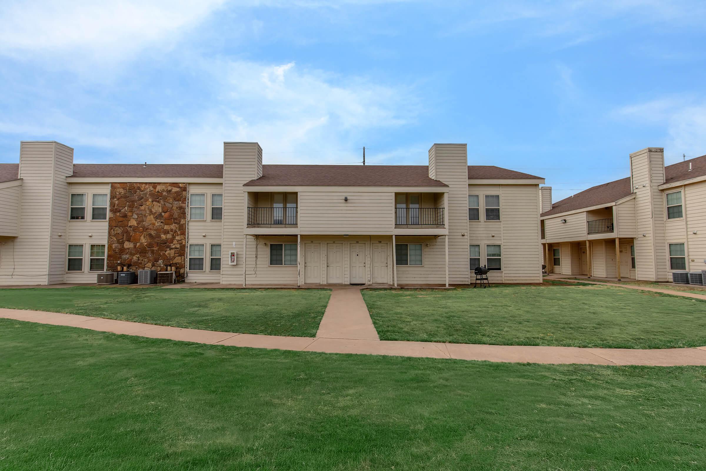 A two-story apartment complex with a light-colored exterior, featuring a stone accent wall. The building has several balconies and is surrounded by well-maintained green grass and a concrete path. The sky is partly cloudy, creating a bright atmosphere.