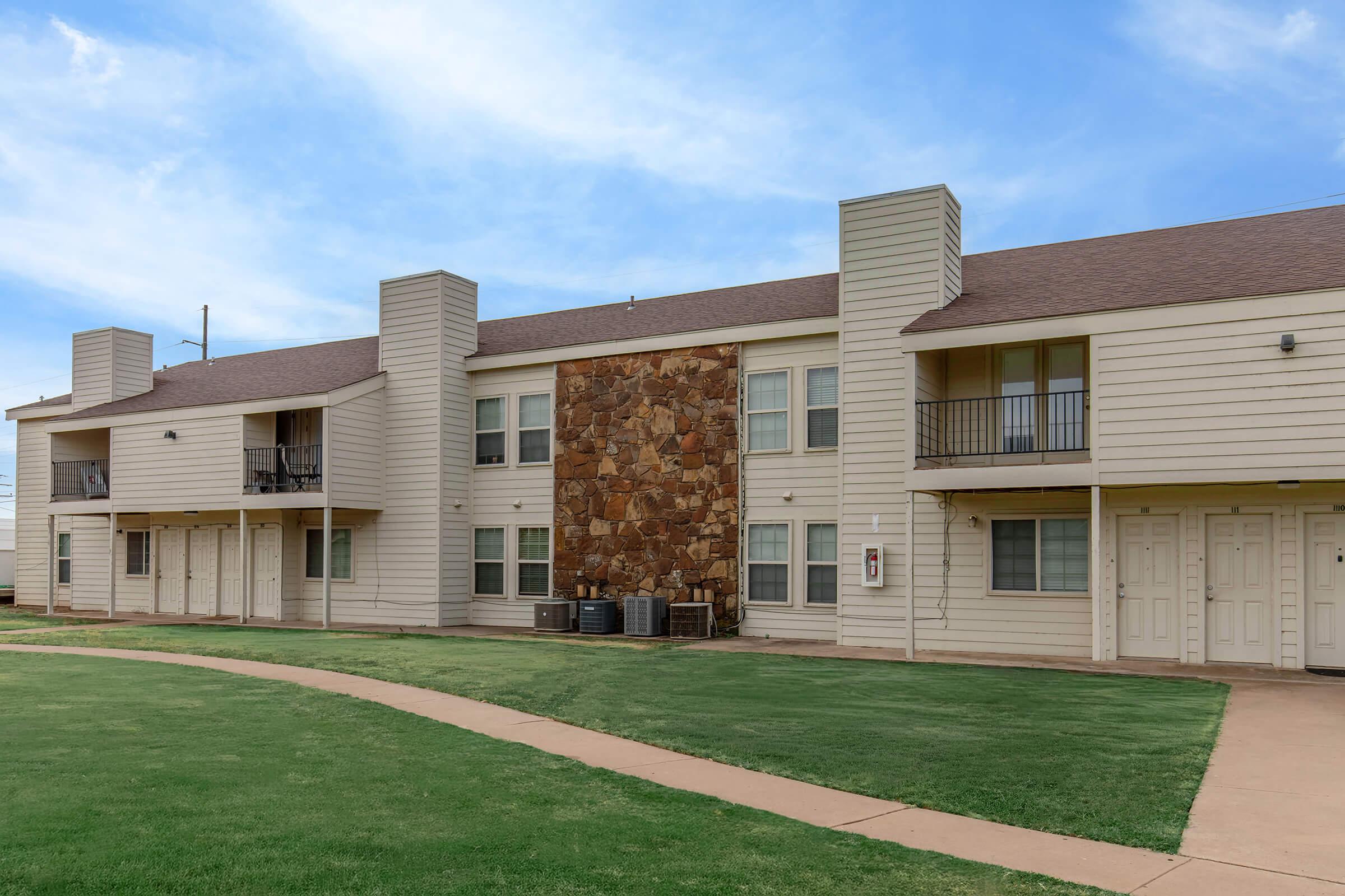 Two-story apartment building with a beige exterior and a rock accent wall. The structure features multiple doors and windows, with air conditioning units on the ground level. Surrounding the building is a well-maintained grassy area and a curved pathway leading to the entrance. The sky is clear and blue.
