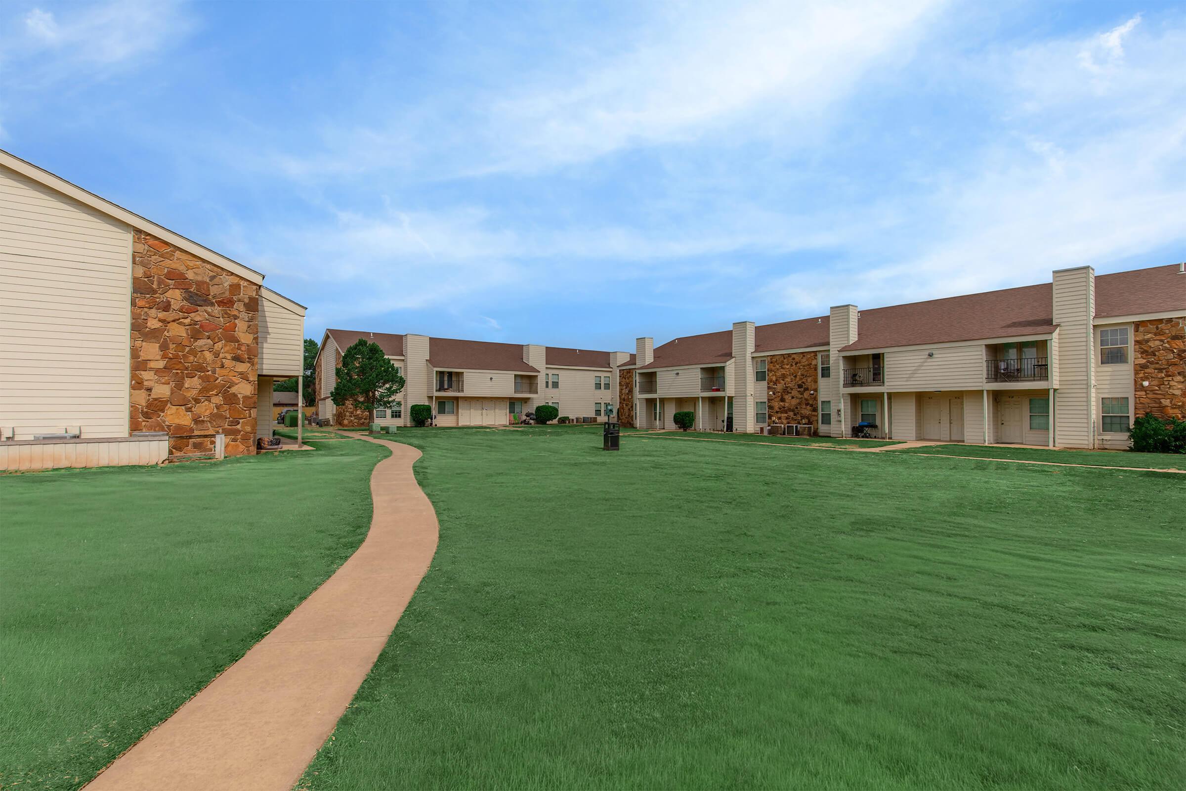 A pathway meanders through a well-maintained grassy area, flanked by two-story apartment buildings with stone and siding exteriors. The sky is blue with a few clouds, creating a pleasant atmosphere. Trees and landscaping enhance the residential setting.