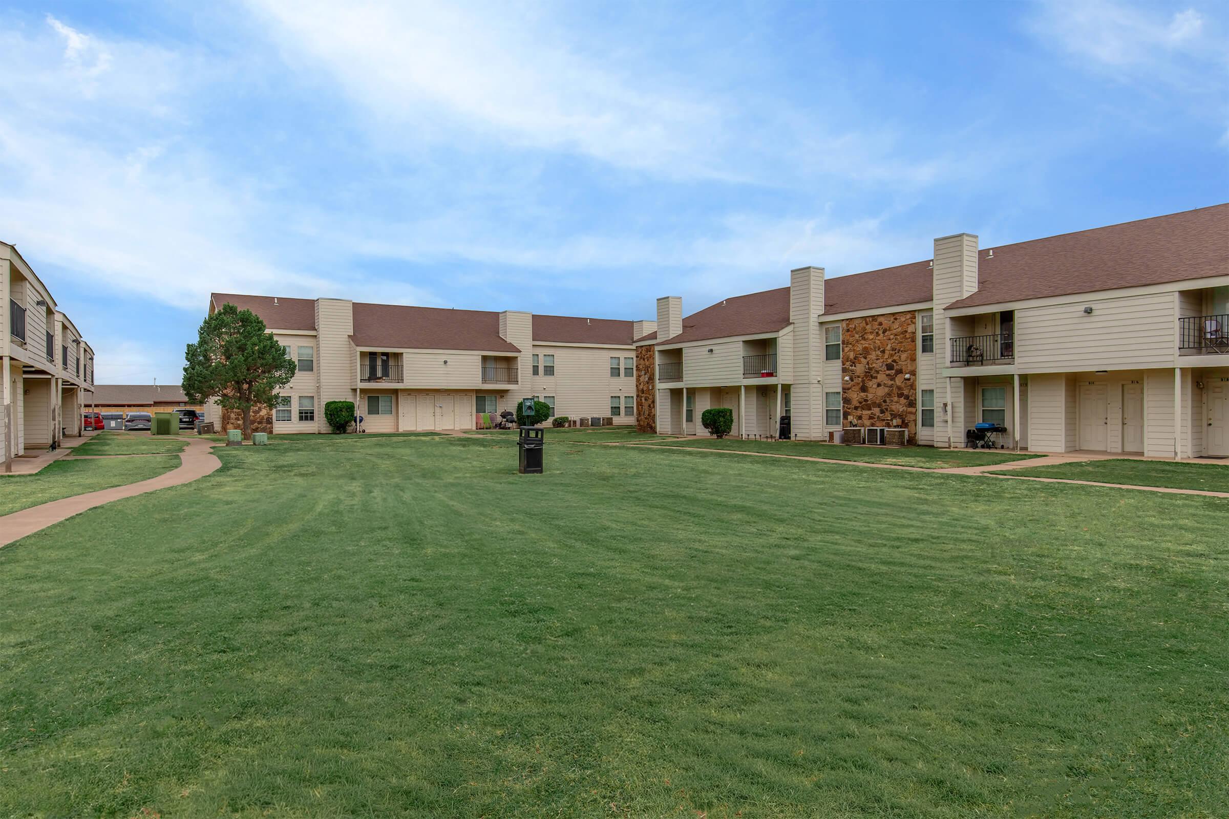 A view of a residential apartment complex with multiple buildings. The two-story structures have a mix of beige siding and stone accents. A green lawn leads to the entrance of the apartments, and there are well-maintained walkways and a few trees in the area, under a blue sky.