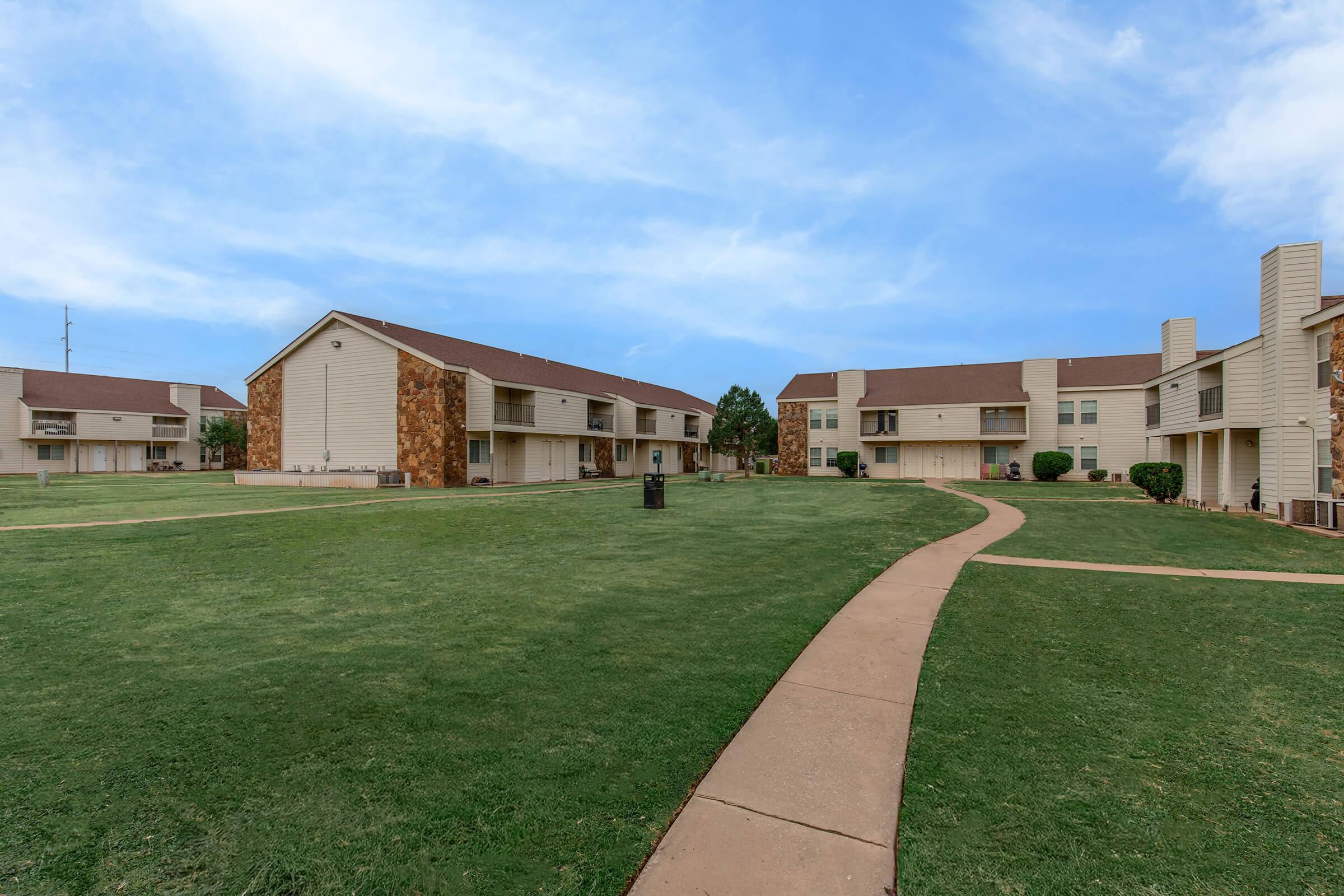 A landscaped residential area featuring two-story apartment buildings with stone accents. A paved pathway winds through a green lawn, with trees and shrubs lining the property. Clear blue sky can be seen in the background, creating a bright and inviting atmosphere.