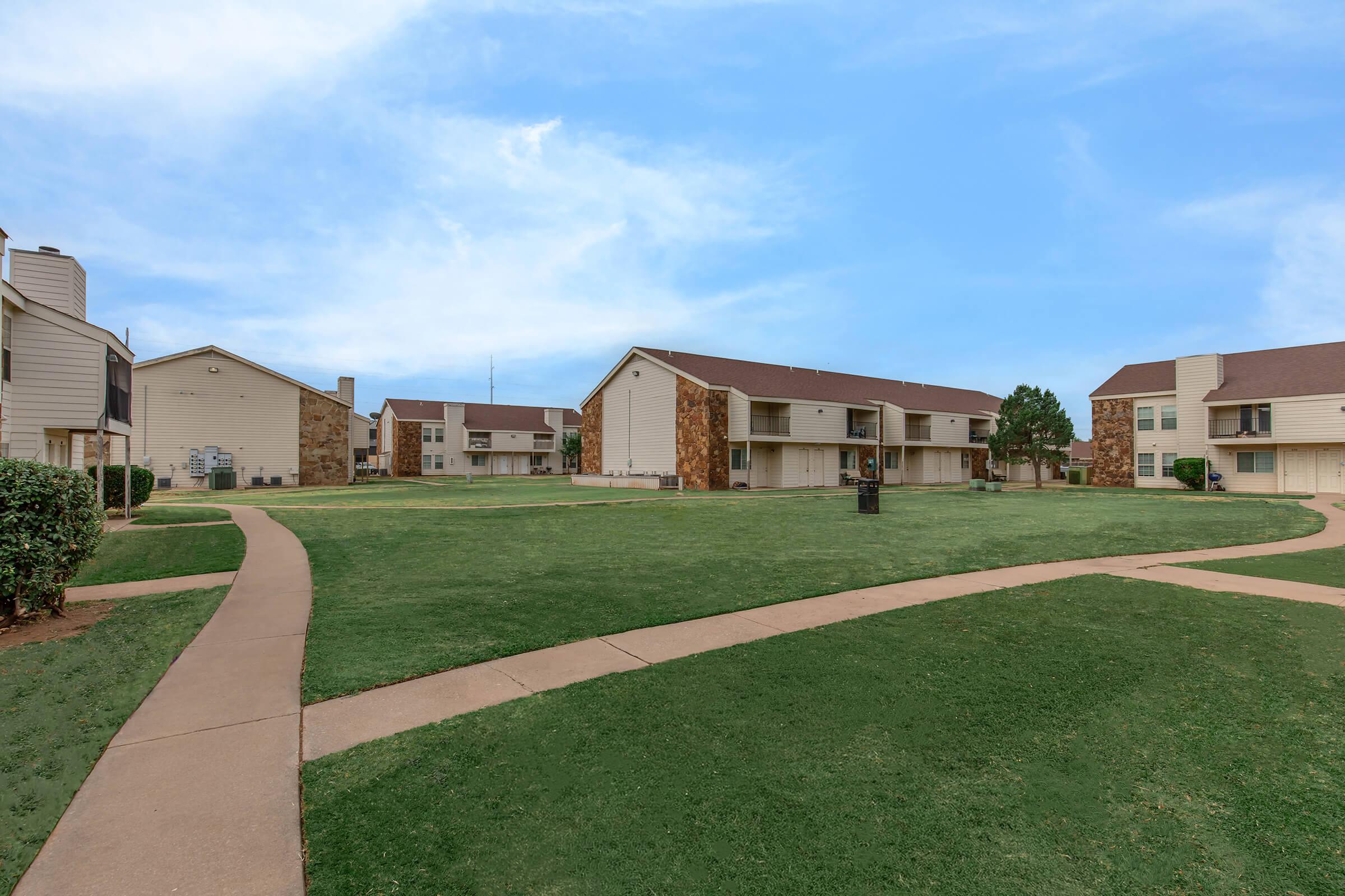 A spacious outdoor area of a residential complex featuring multiple two-story buildings with light-colored siding and stone accents. There are neatly trimmed lawns and walking pathways, creating a tranquil environment under a blue sky.