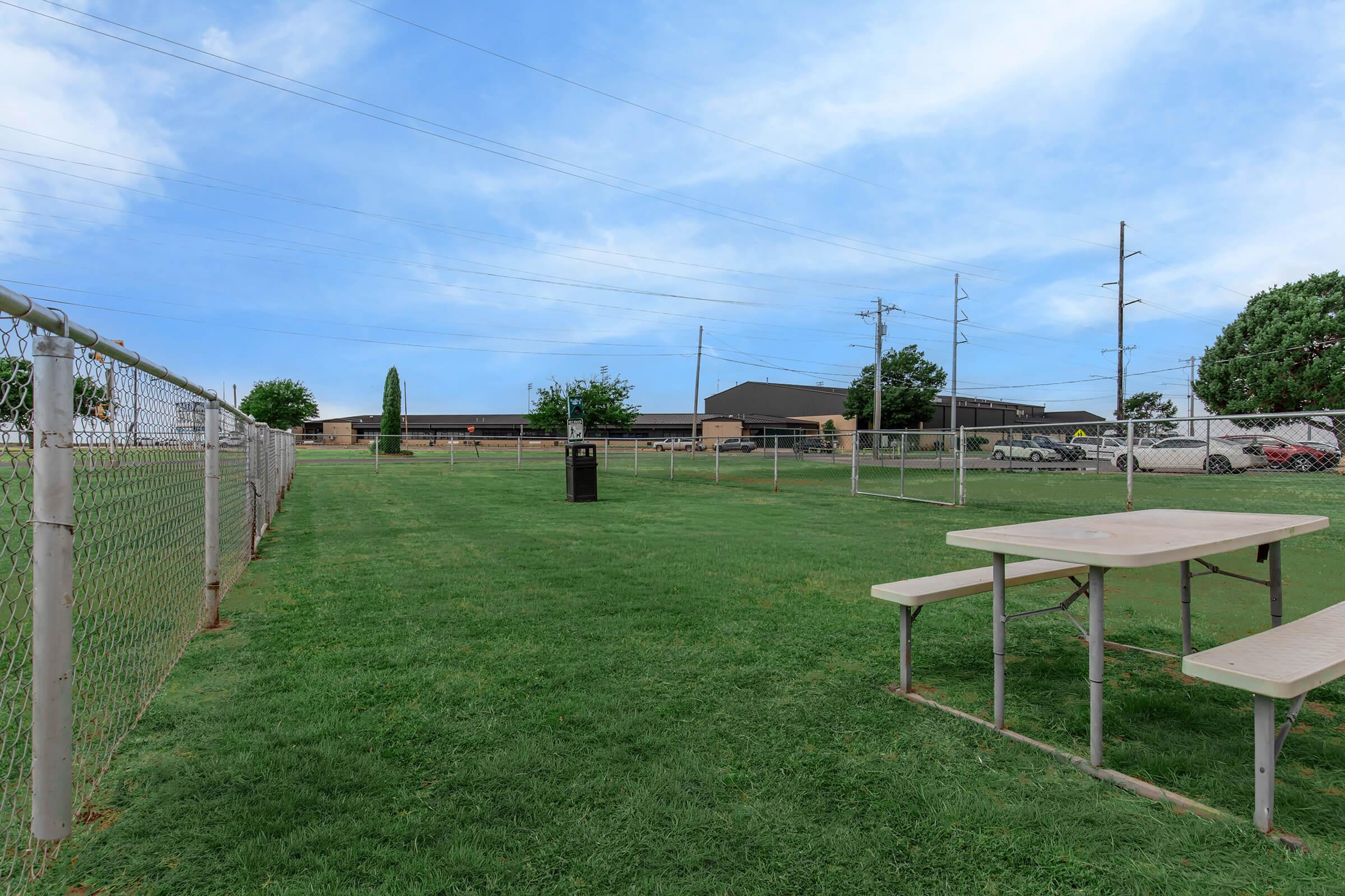 A grassy open area enclosed by a chain-link fence, featuring picnic tables and a trash can. In the background, there are utility poles and a building with a parking area. The sky is clear with a few clouds.