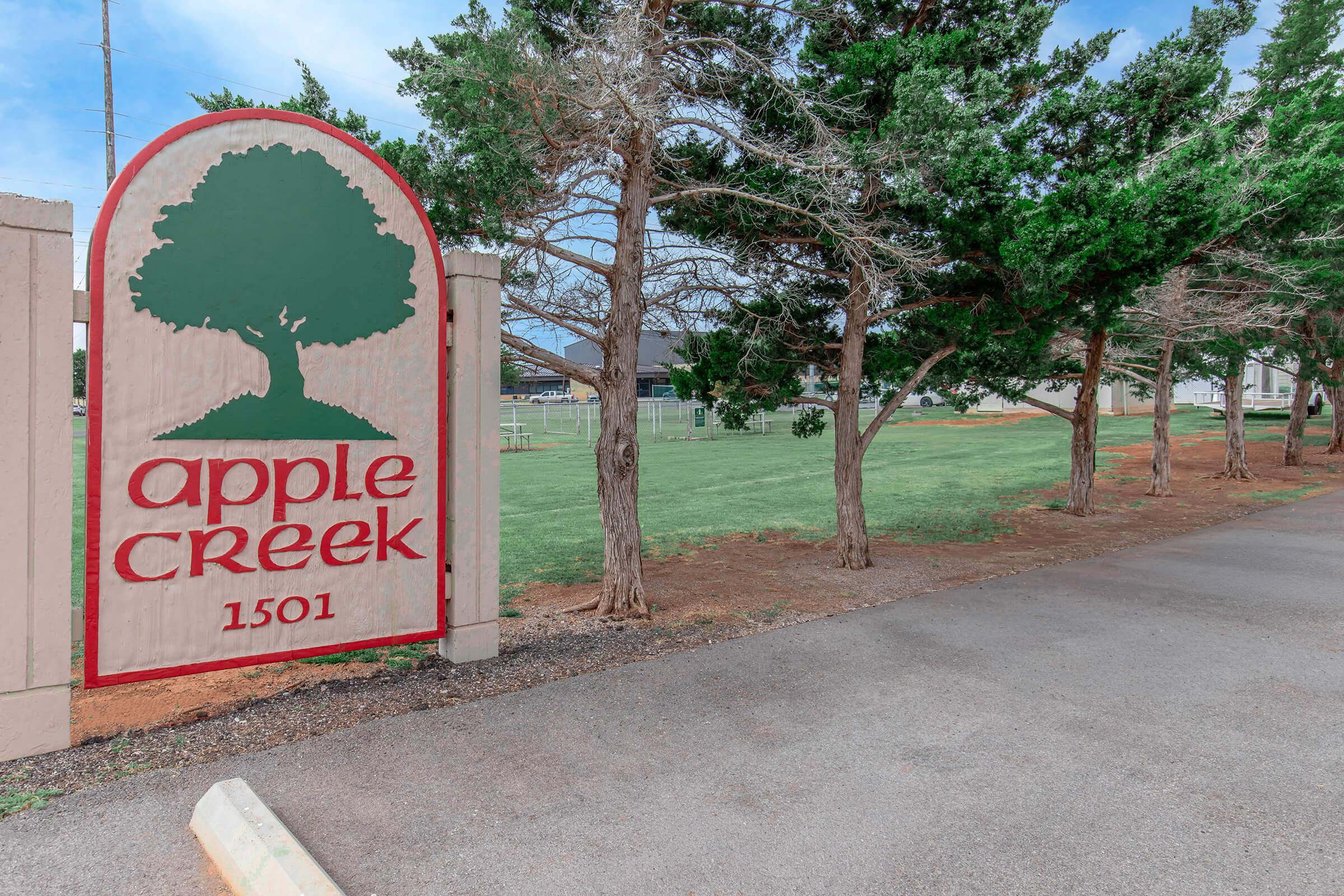 Sign for Apple Creek with a tree logo, displaying the address 1501. Surrounding the sign are neatly trimmed trees and a grassy area, indicating a well-maintained outdoor space. The path leading up to the sign is paved.